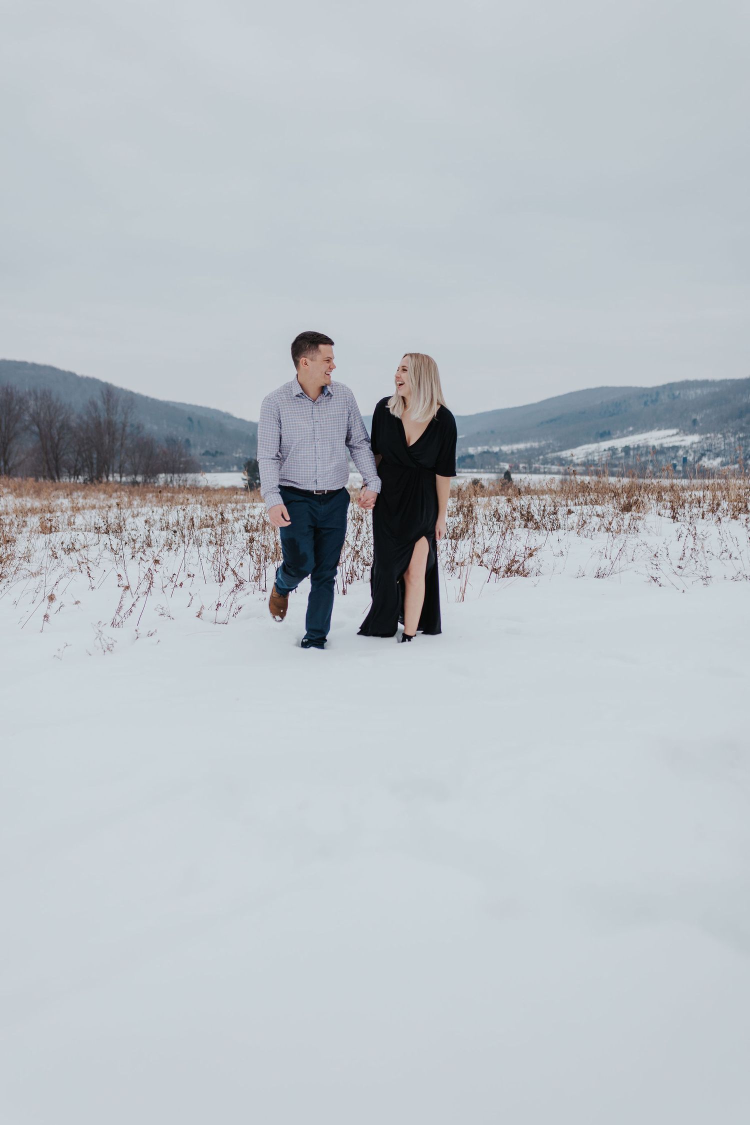 Couple holding hands walking through snowy winter field, woman in black dress and man in gray shirt, mountains behind.