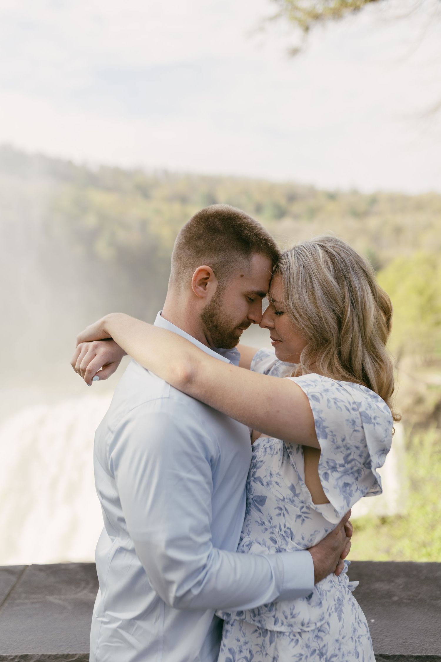 Romantic couple embracing with foreheads touching in front of a misty waterfall backdrop.
