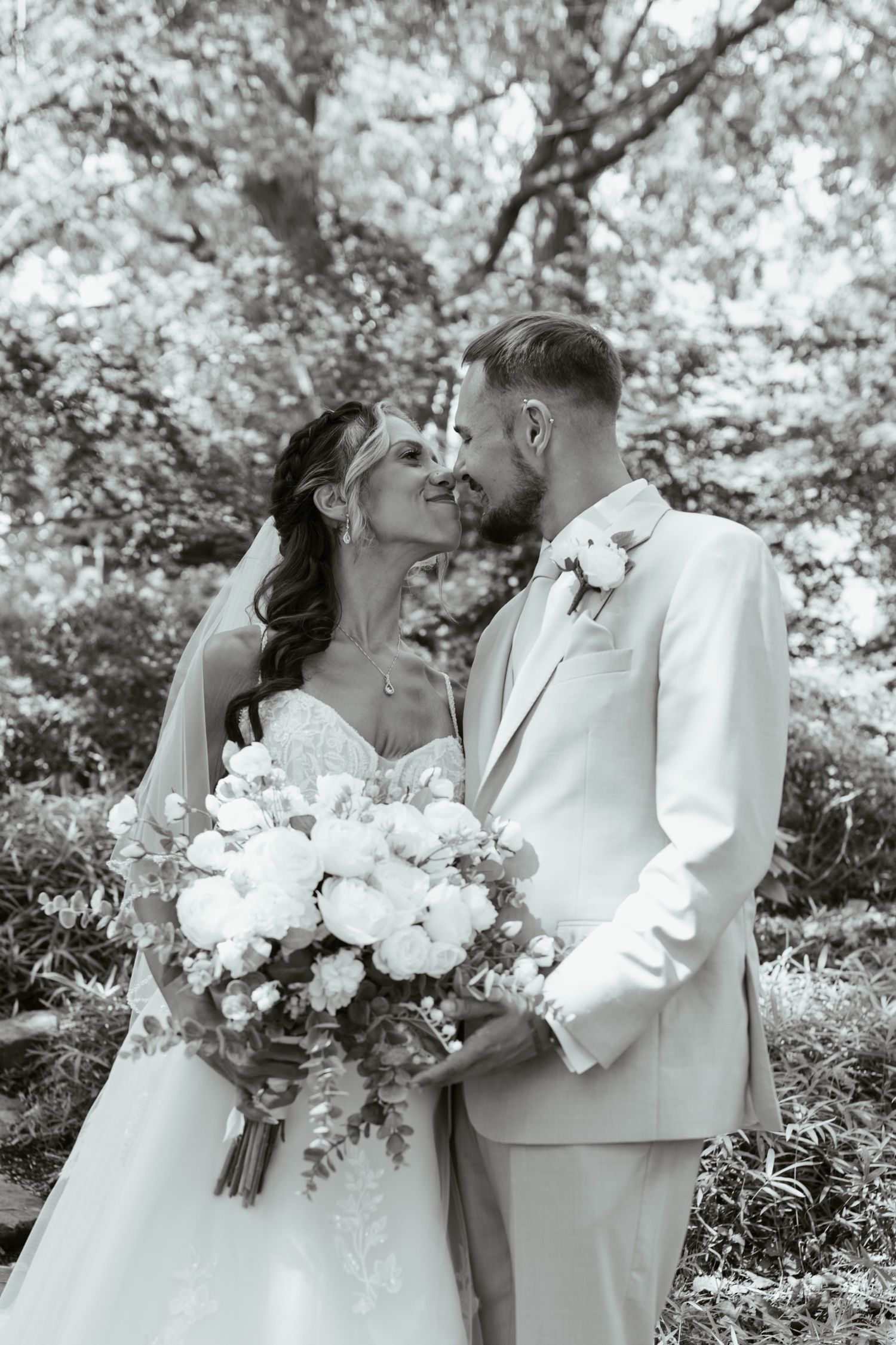 Black and white wedding photo of bride holding bouquet and groom in white suit sharing a romantic moment outdoors.