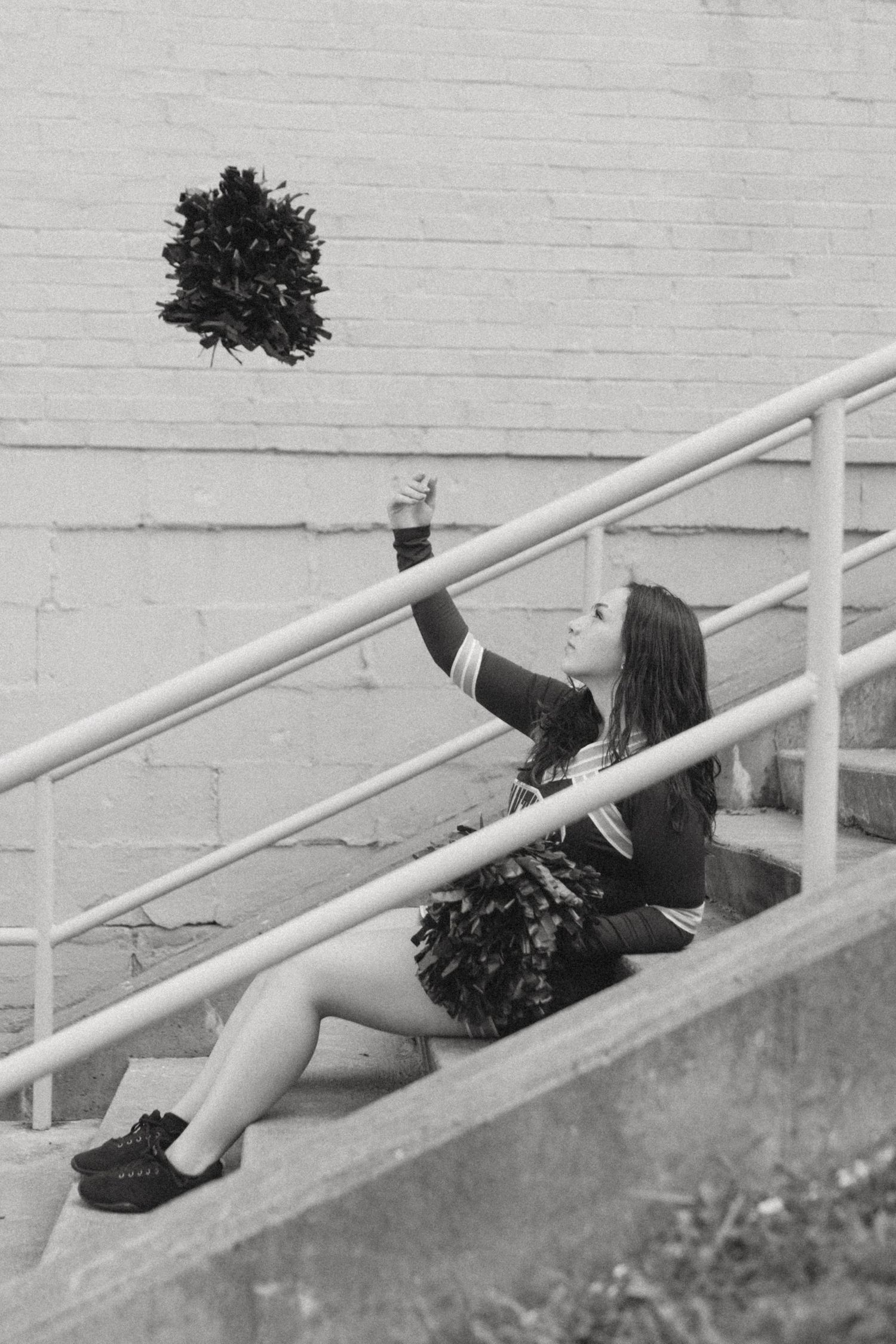 Black and white photo of a cheerleader sitting on stairs tossing a pom-pom in the air.