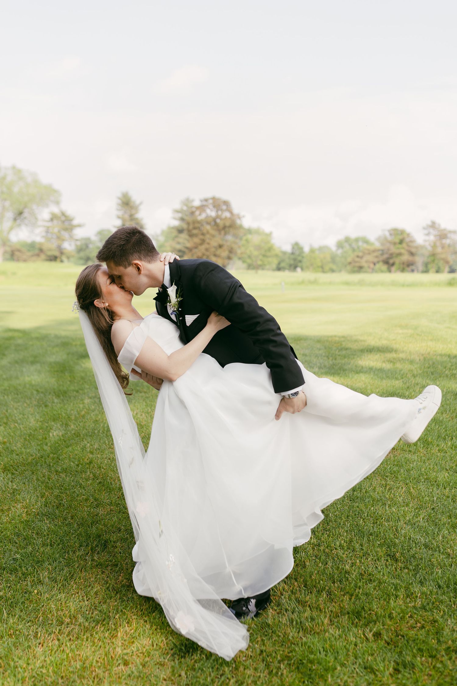 A romantic dip kiss between newlyweds on a sunlit grassy field.