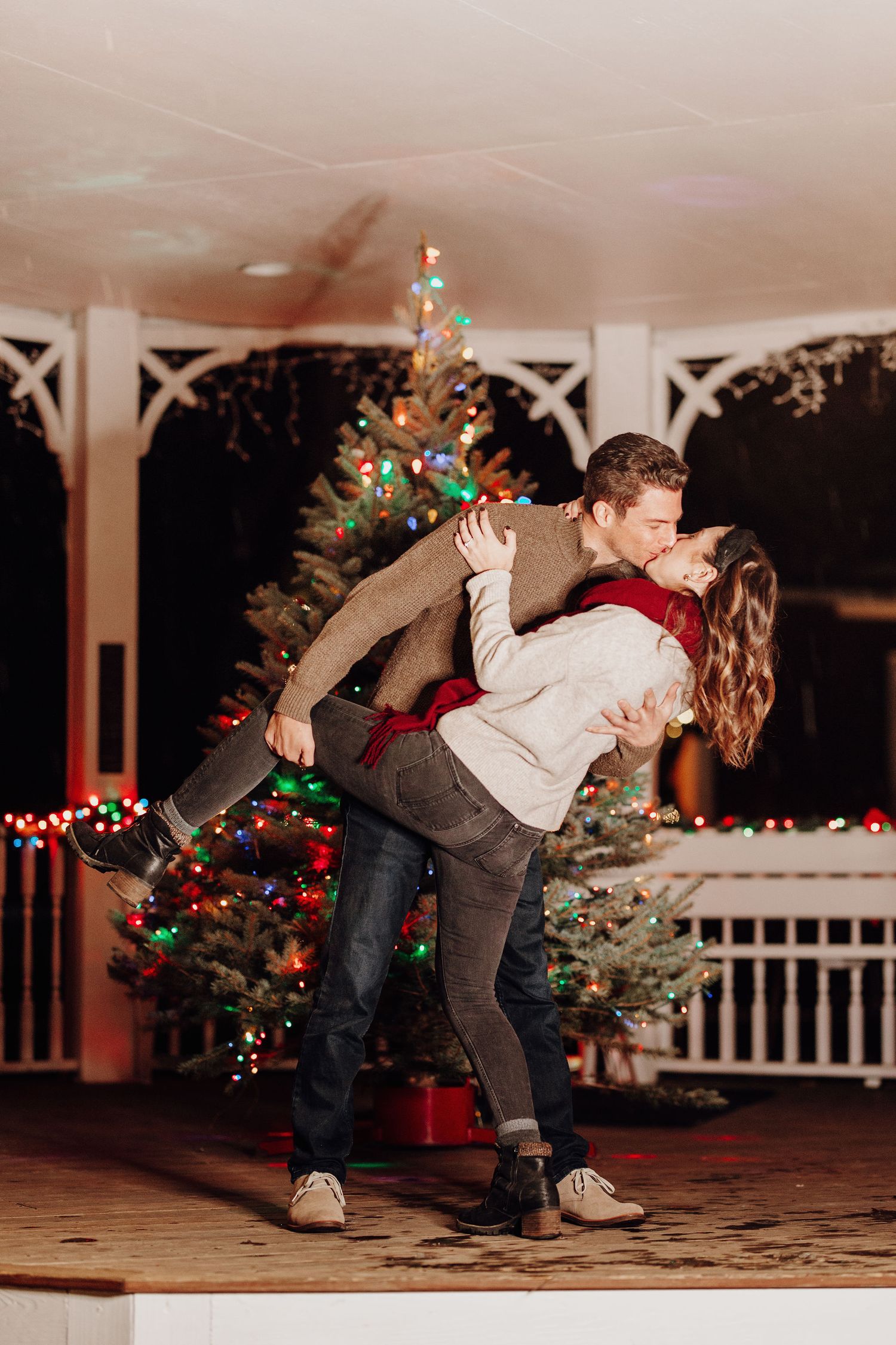 Couple sharing a romantic kiss outdoors near a decorated Christmas tree with colorful lights at night.