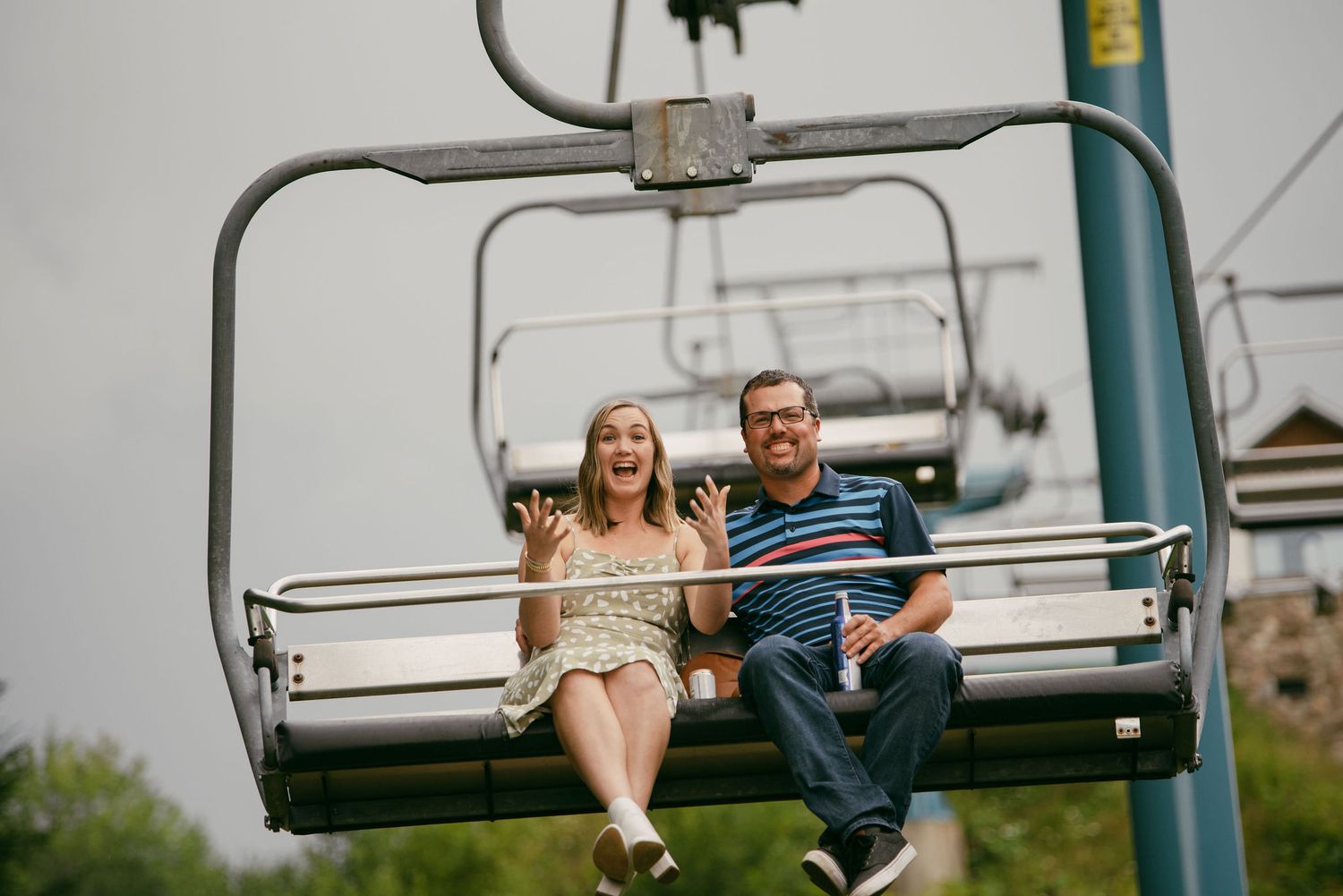 Couple laughing and waving on a ski lift chair during summer at a mountain resort.