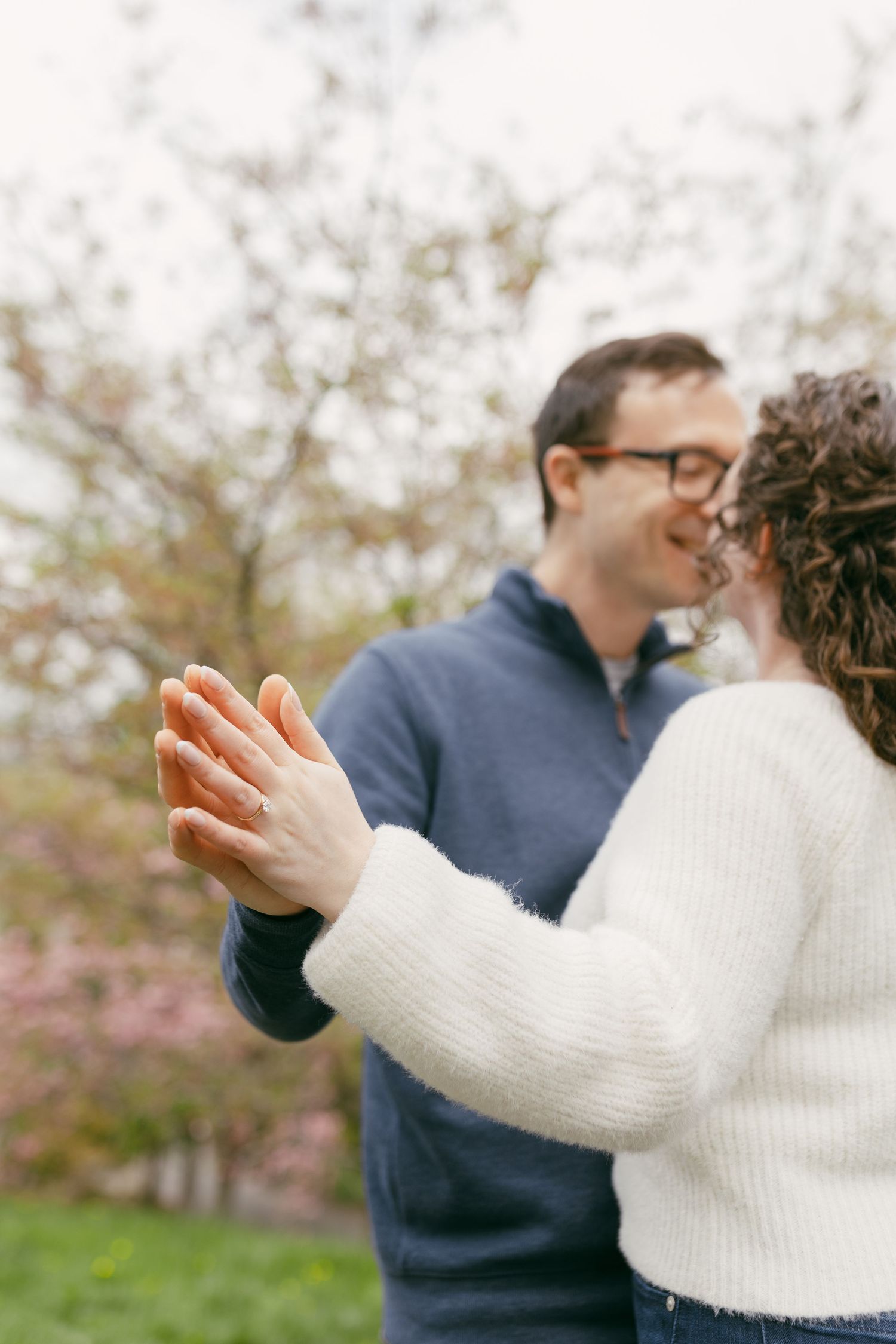 Couple holding hands with engagement ring visible, smiling outdoors near blooming trees in spring.