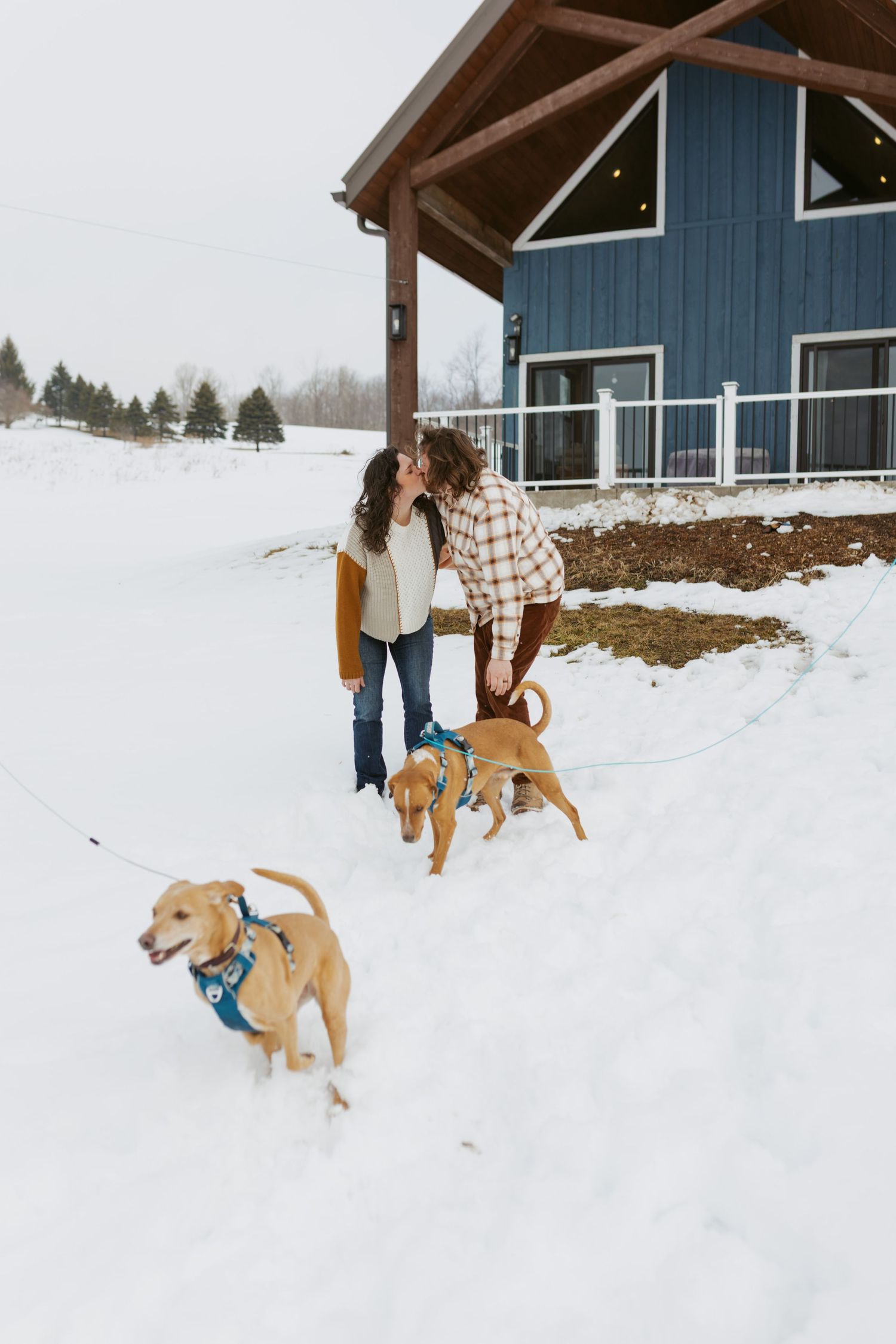 Couple sharing a kiss in the snow with two golden dogs on leashes outside a blue A-frame house.