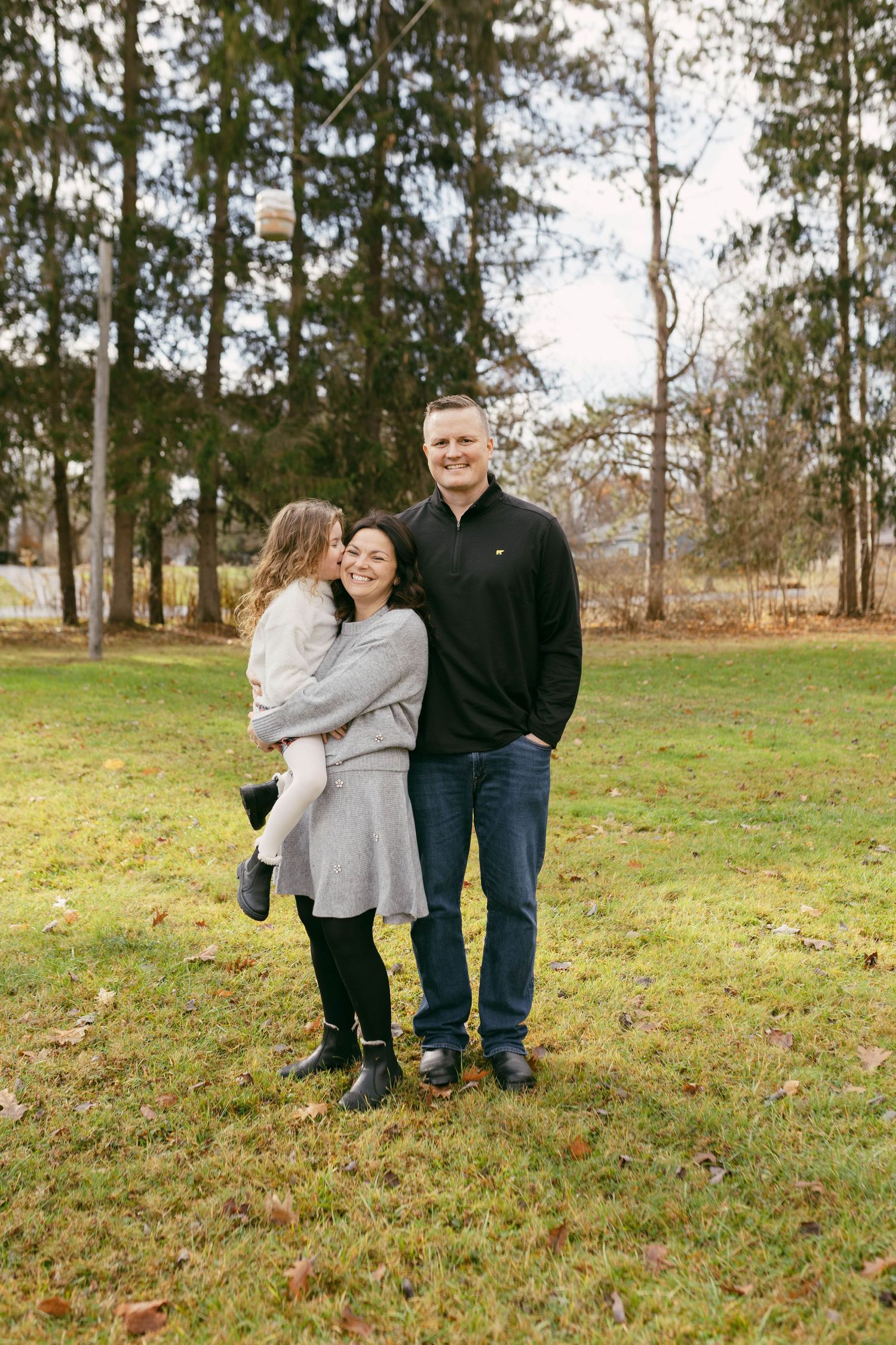 A smiling family of three poses outdoors on a grassy lawn surrounded by tall pine trees during autumn.