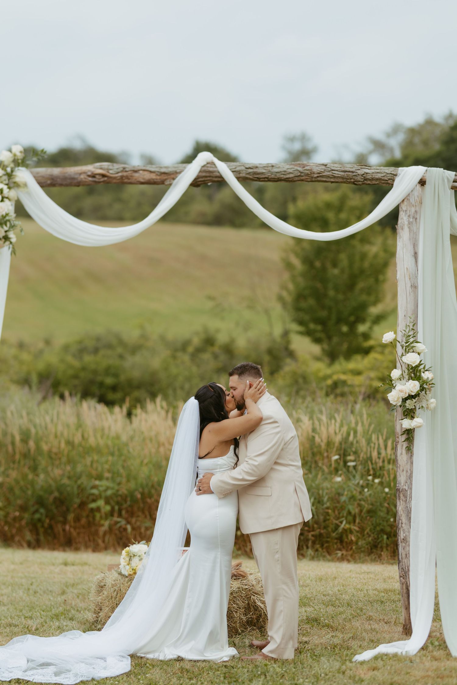 Bride and groom share first kiss under rustic wooden arch with white draping and flowers at outdoor wedding ceremony.