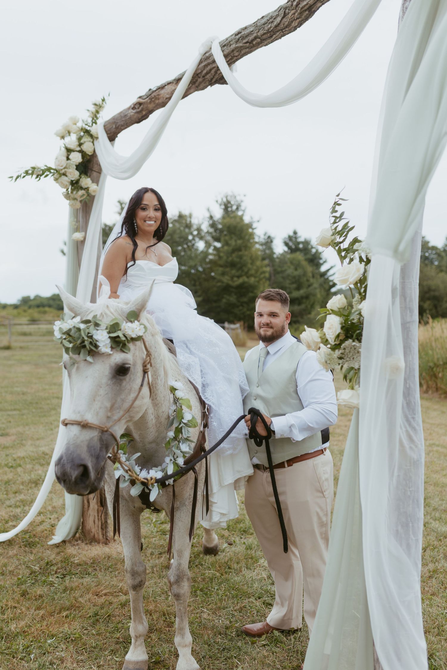 Bride in white gown sits on decorated white horse as groom stands beside her under floral arch at outdoor wedding.