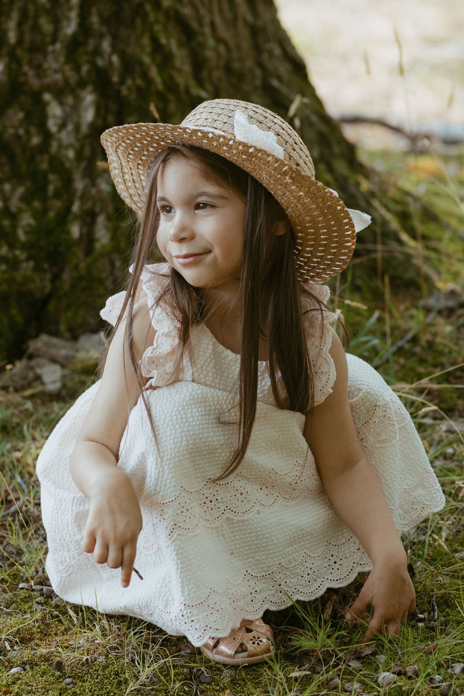Young girl in white lace dress and straw hat sitting by tree trunk in nature, smiling outdoors.