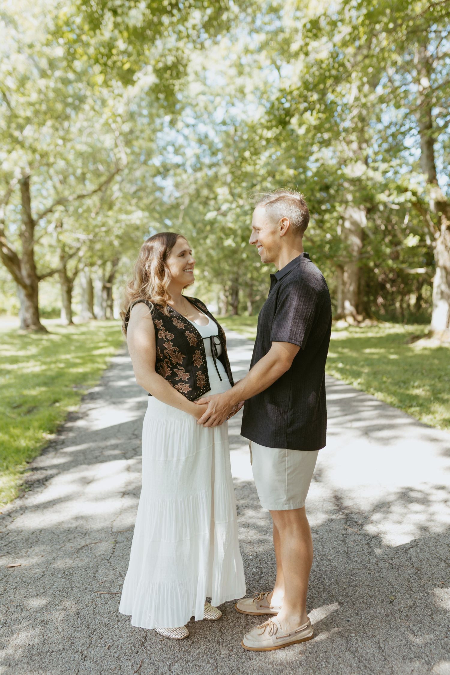 Couple holding hands and smiling at each other on a sunny tree-lined path, woman in white dress, man in black shirt.