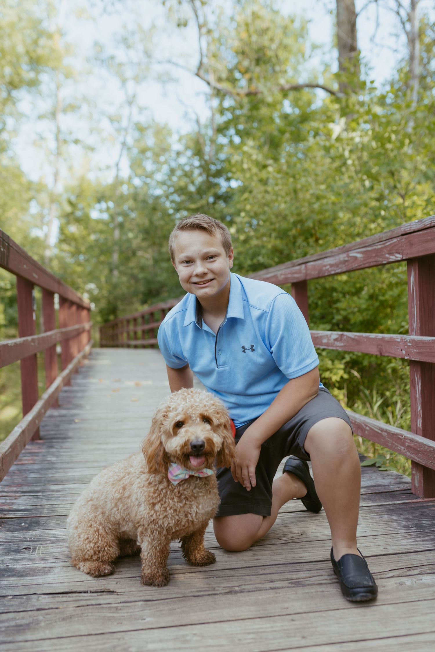 A smiling boy in a blue polo shirt kneels beside a fluffy golden doodle dog on a wooden bridge surrounded by trees.