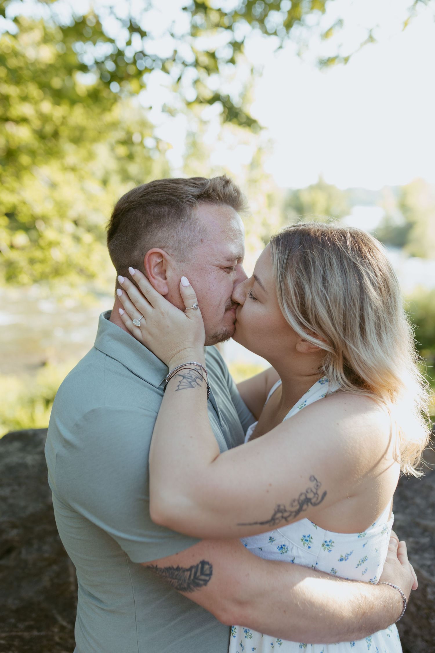 A couple shares a romantic kiss outdoors, with the woman wearing an engagement ring and both displaying tattoos.
