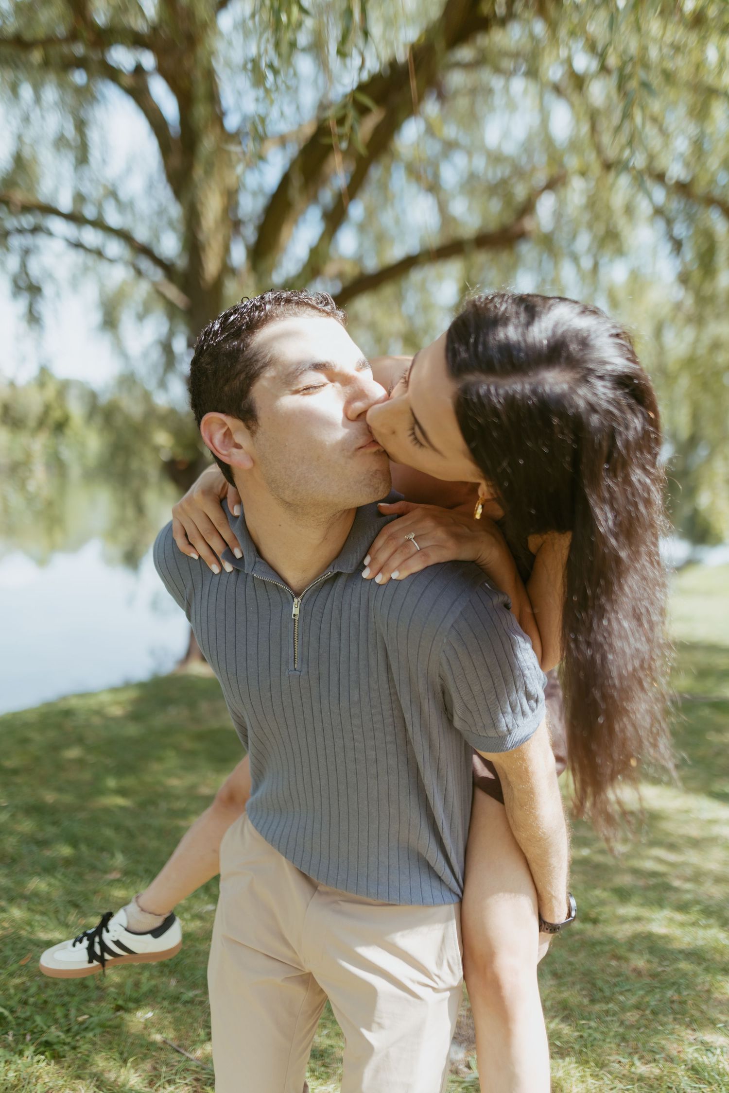 Couple sharing a romantic piggyback kiss outdoors under a willow tree near a lake on a sunny day.