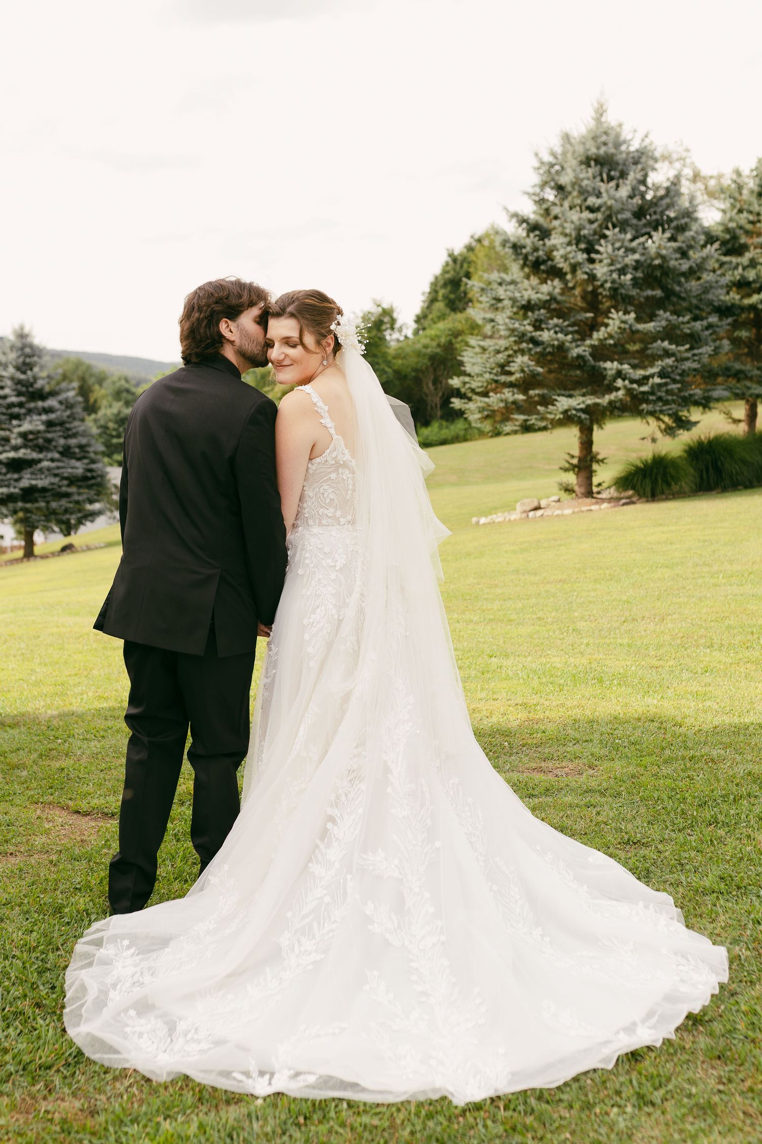 A romantic back view of newlyweds embracing on a grassy hillside with evergreen trees.