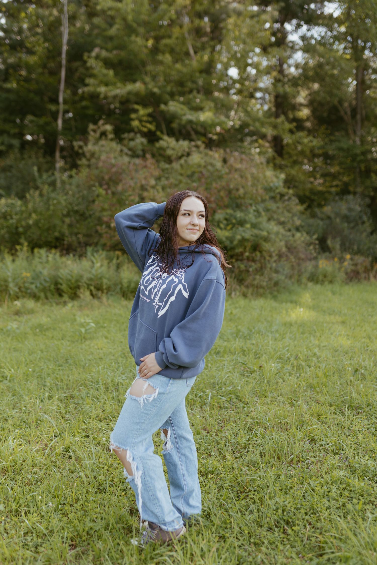 Young woman in blue hoodie and ripped jeans posing in a green field with trees in the background.