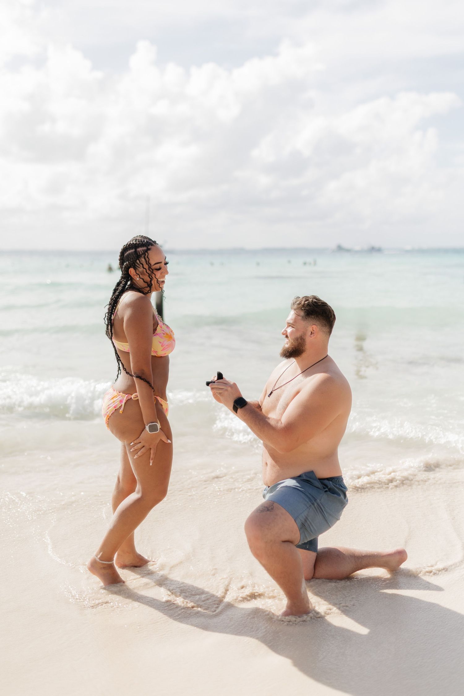 Man proposes to woman on a tropical beach, kneeling in the sand with a ring box while she stands in a bikini.