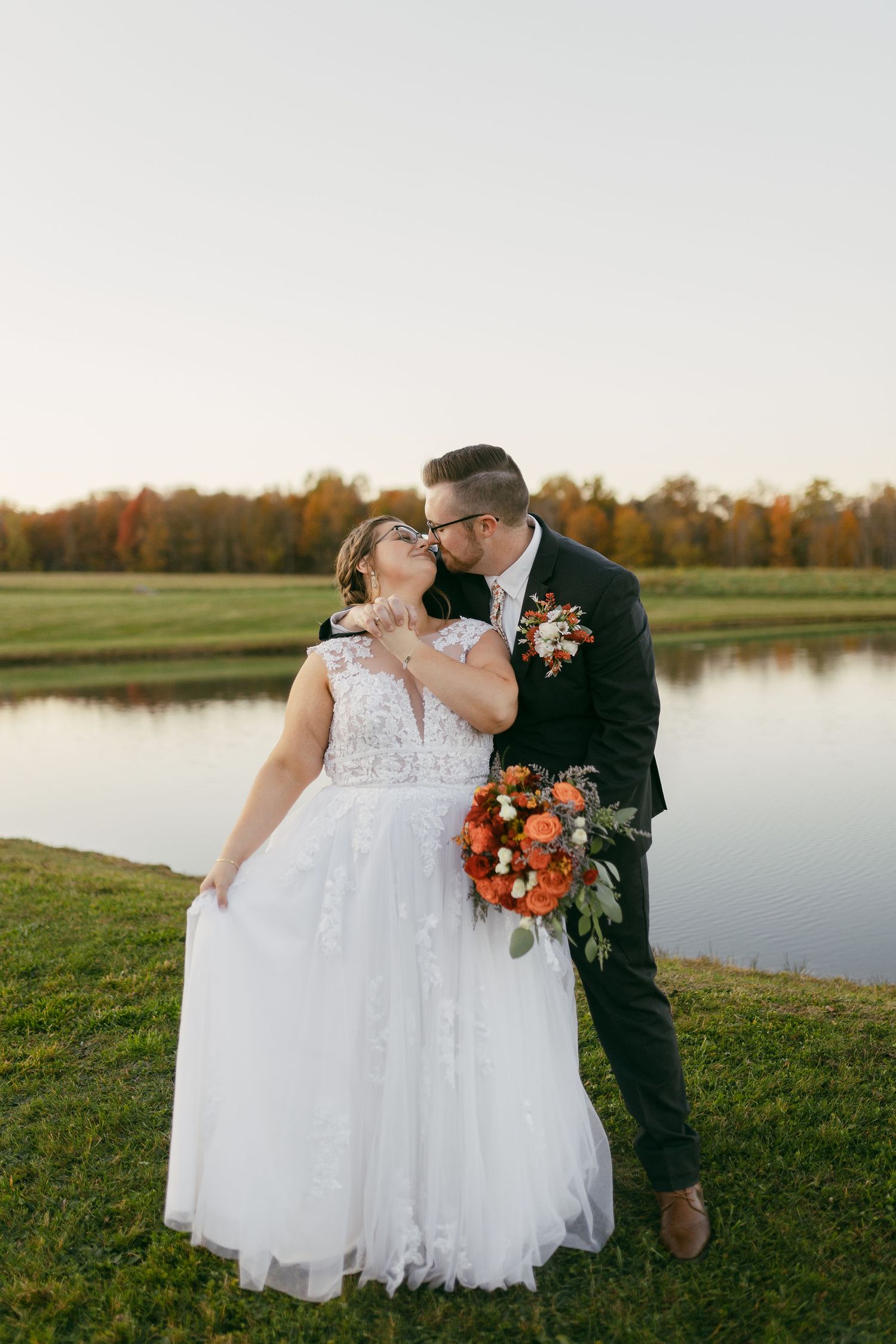 Wedding couple sharing intimate moments by a serene lake during autumn sunset.