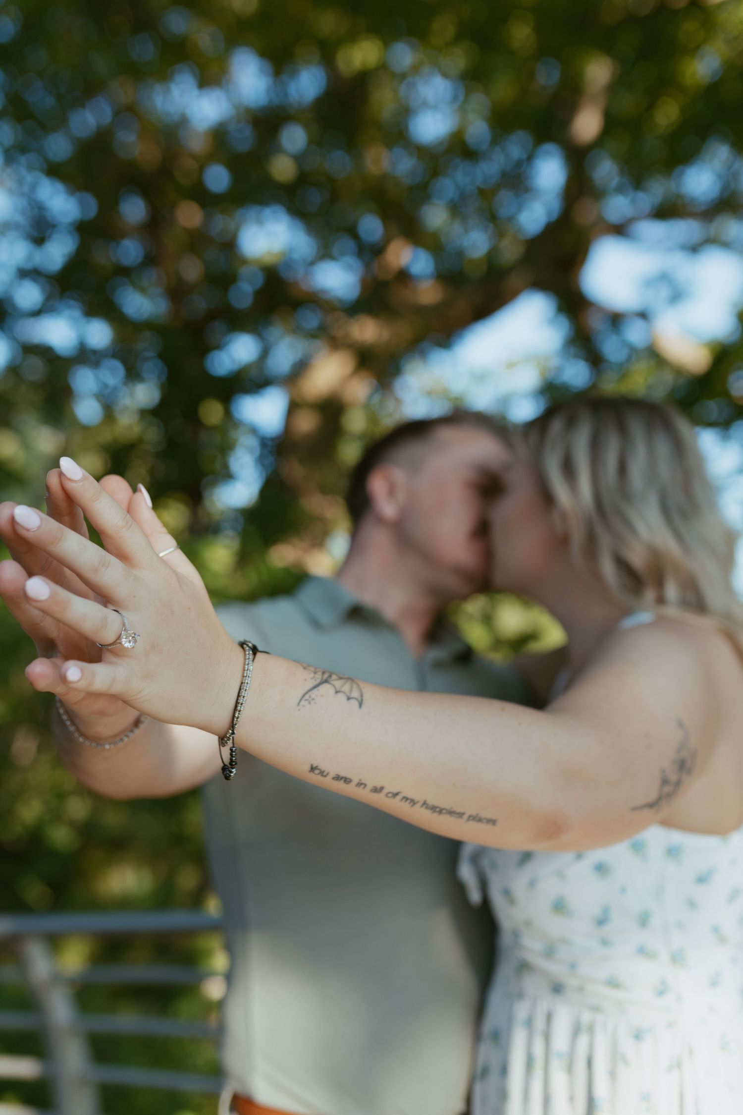 Couple kissing while showing off engagement ring with tattoos visible on womans arm.