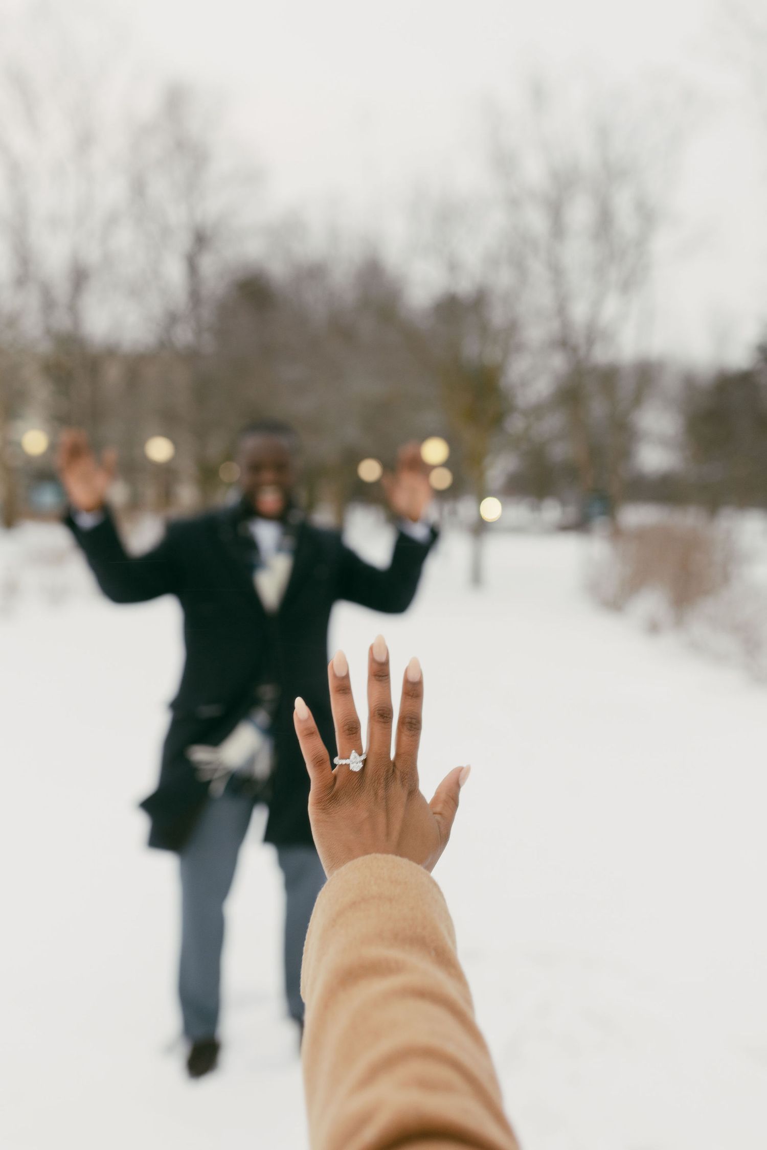 Newly engaged woman shows off diamond ring in snowy park with excited partner celebrating in background.