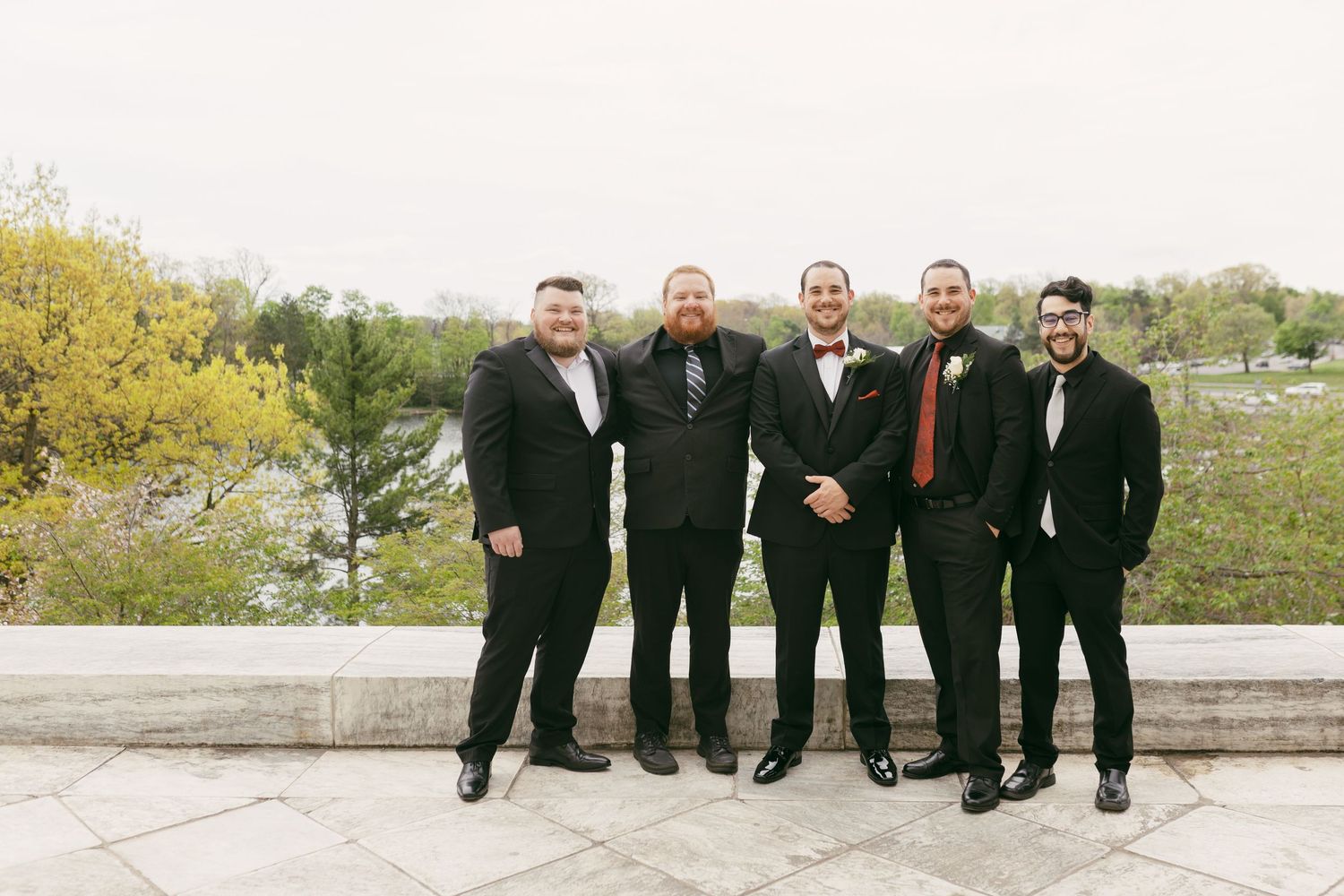 Groom and groomsmen in black suits posing outdoors with scenic park backdrop on wedding day.