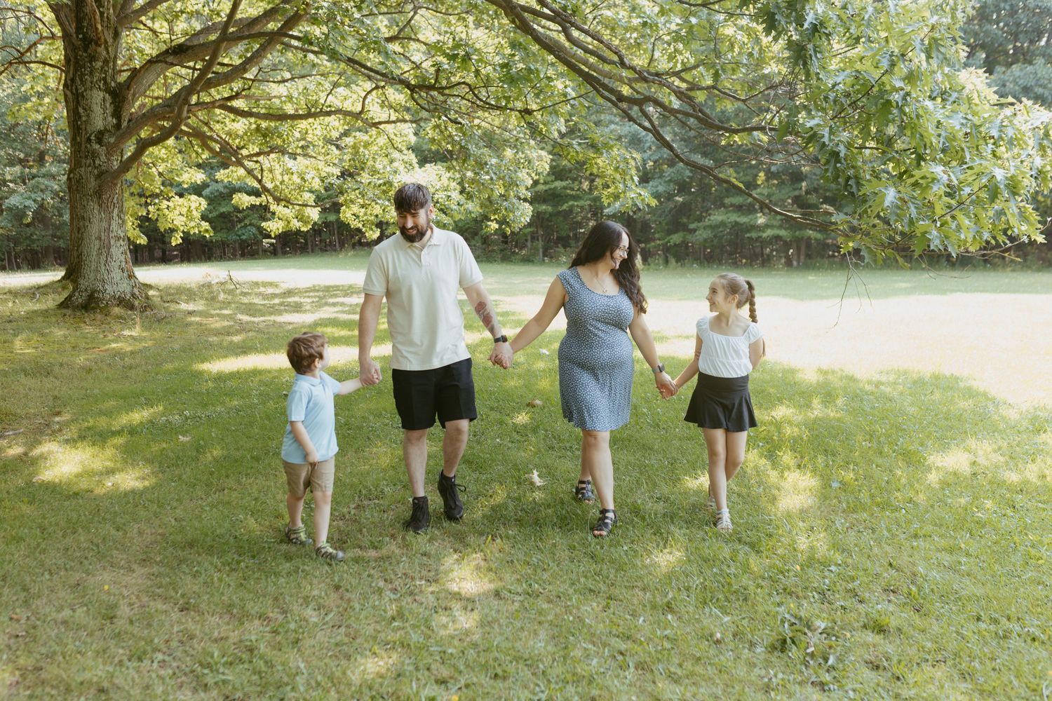 Family of four holding hands walking in a sunny green park under large trees.