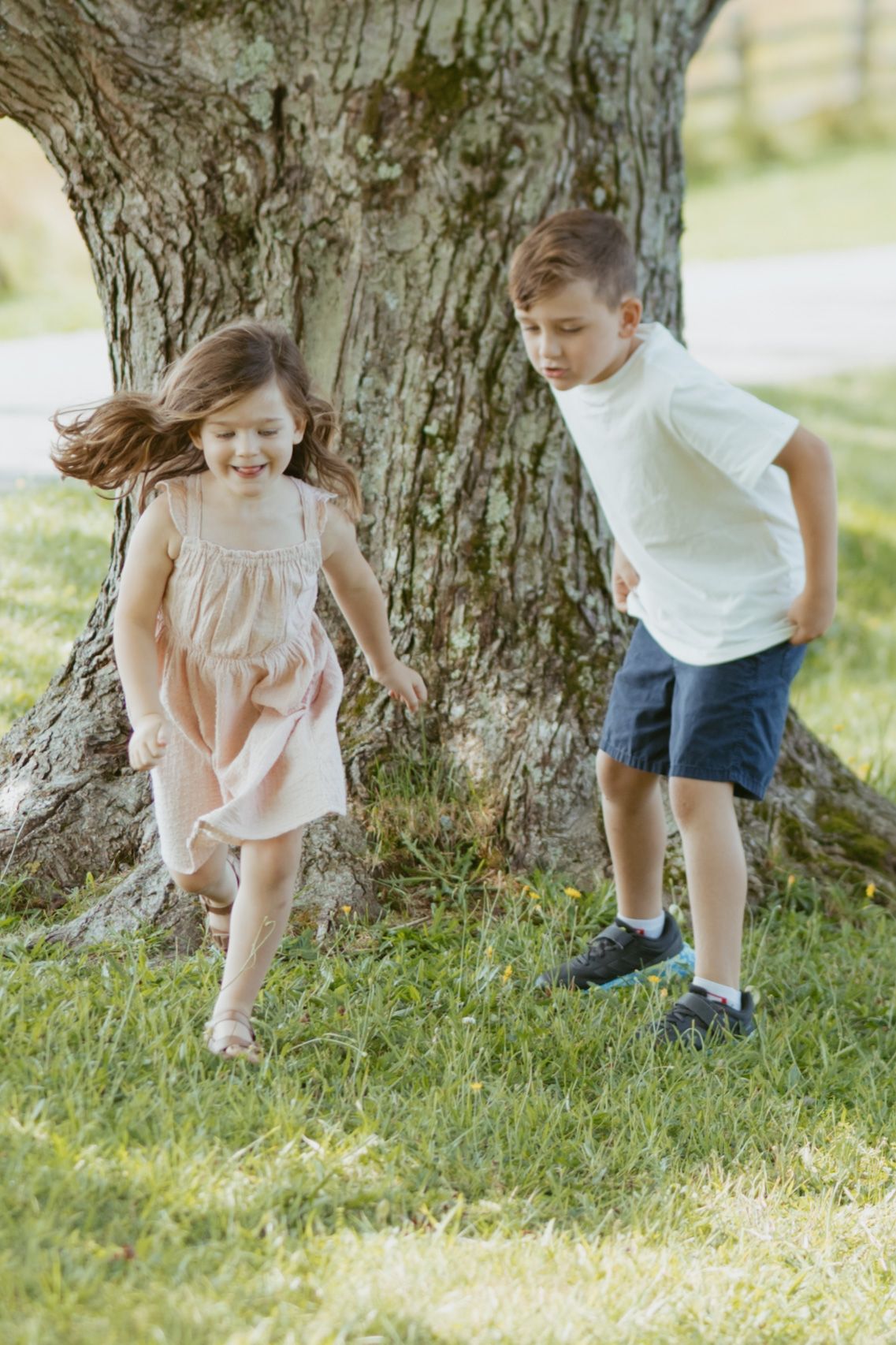 Two children playing and running on green grass near a large tree trunk on a sunny day.