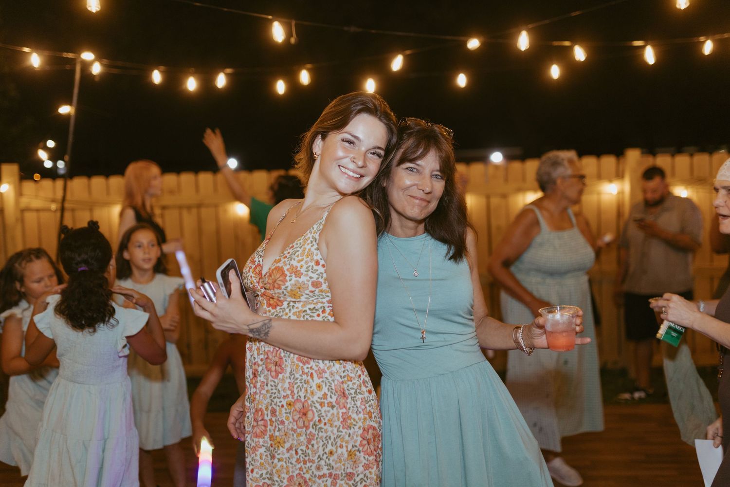 Two women smiling and posing together at an outdoor evening party with string lights and guests dancing.