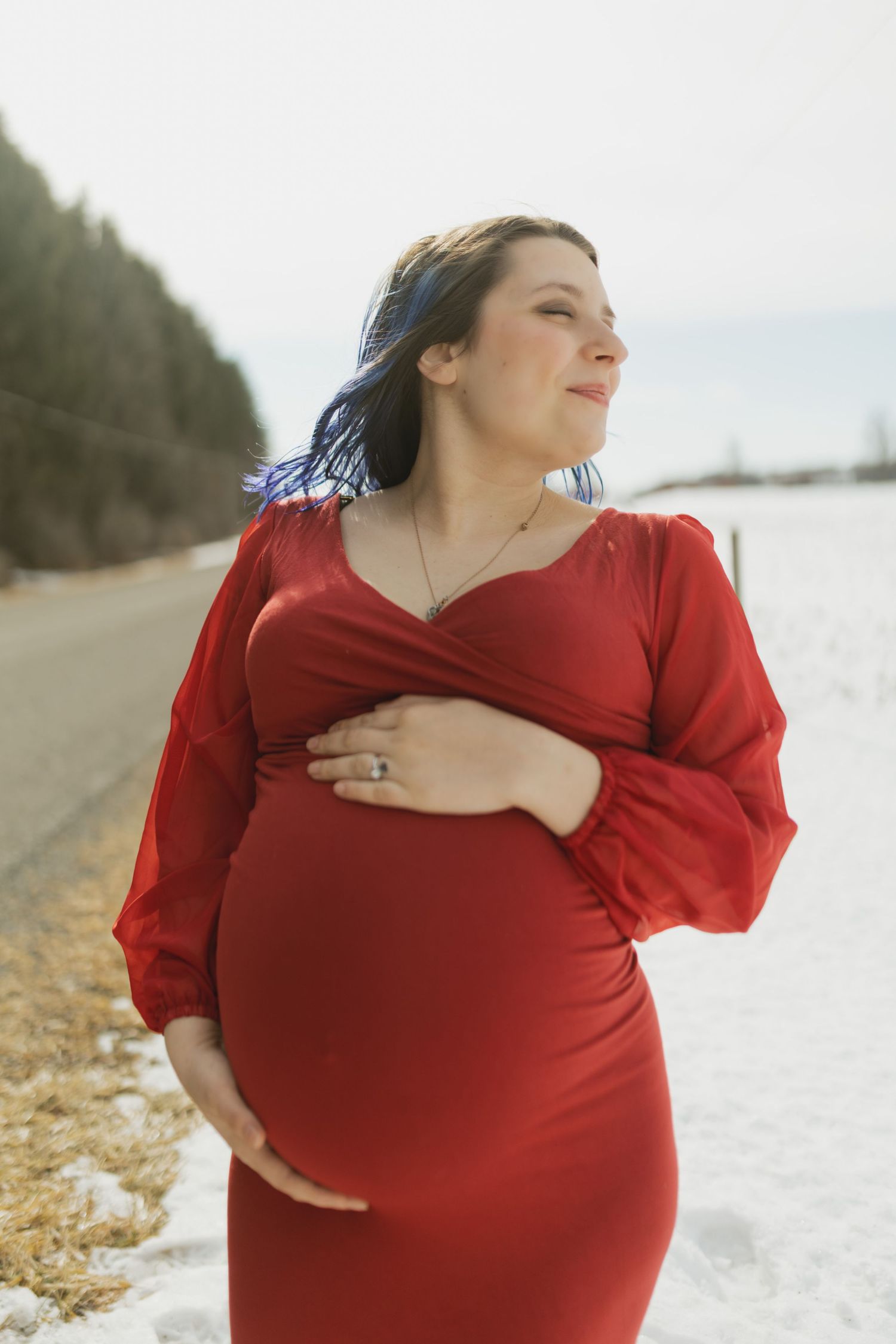 Pregnant woman in red wrap dress holding baby bump outdoors in winter, smiling serenely beside a snowy road.