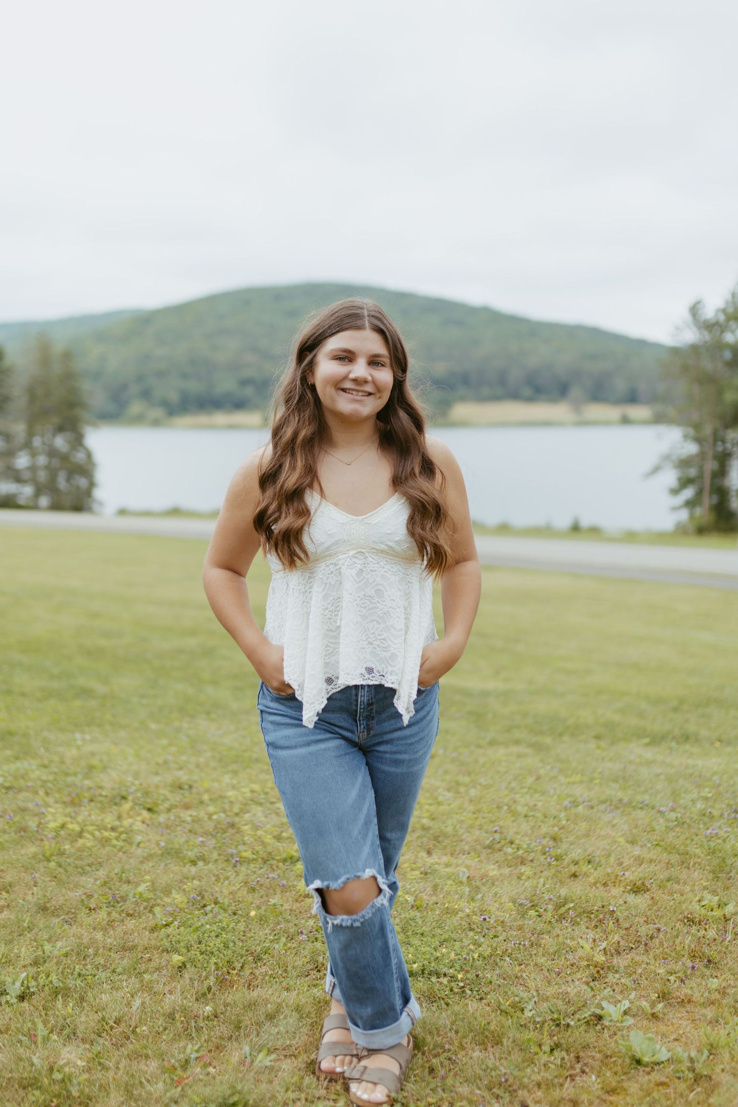 Young woman in white lace tank top and ripped jeans smiling outdoors near a lake with green hills behind her.