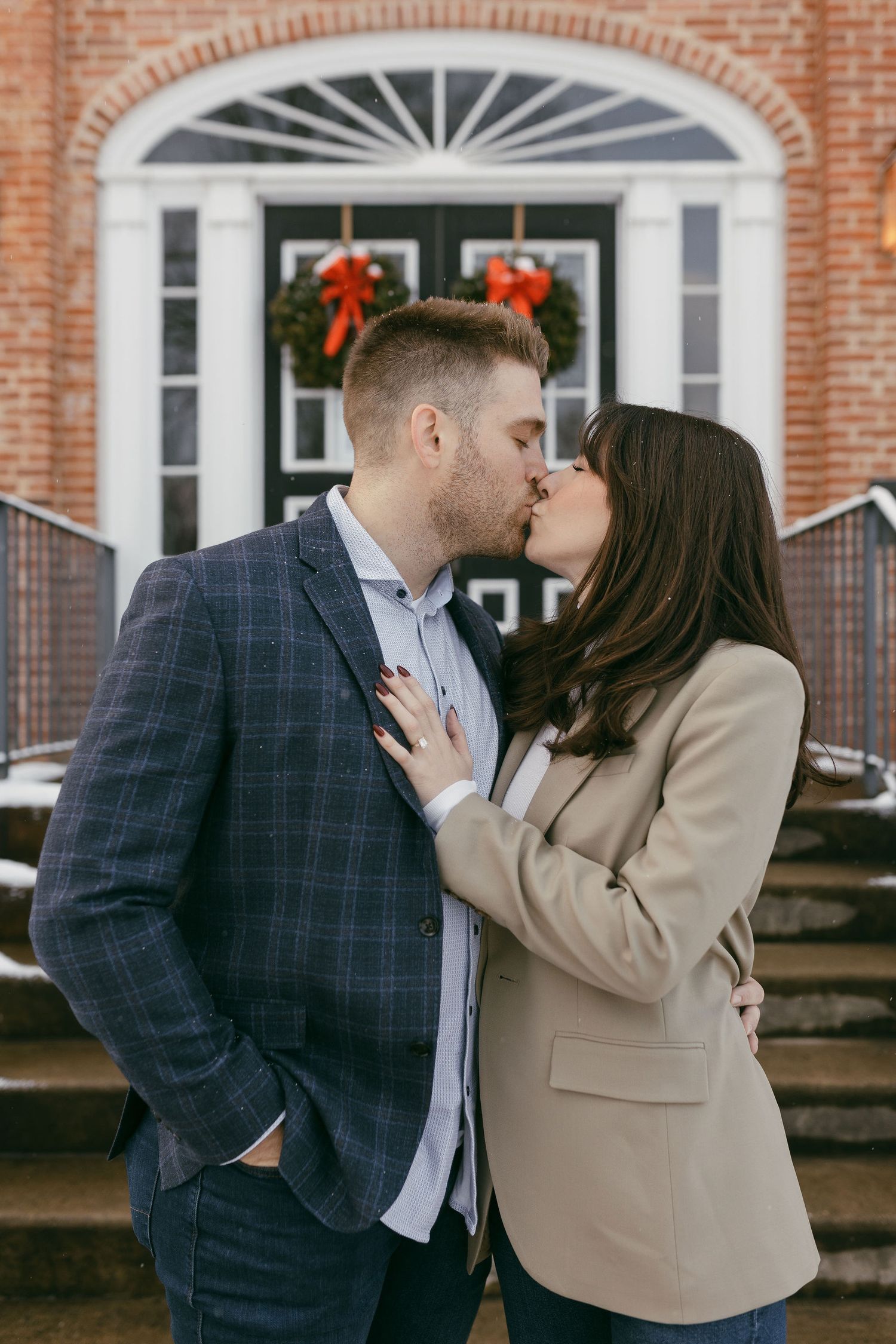 Couple sharing a romantic kiss in front of a brick building with Christmas wreaths on the door.