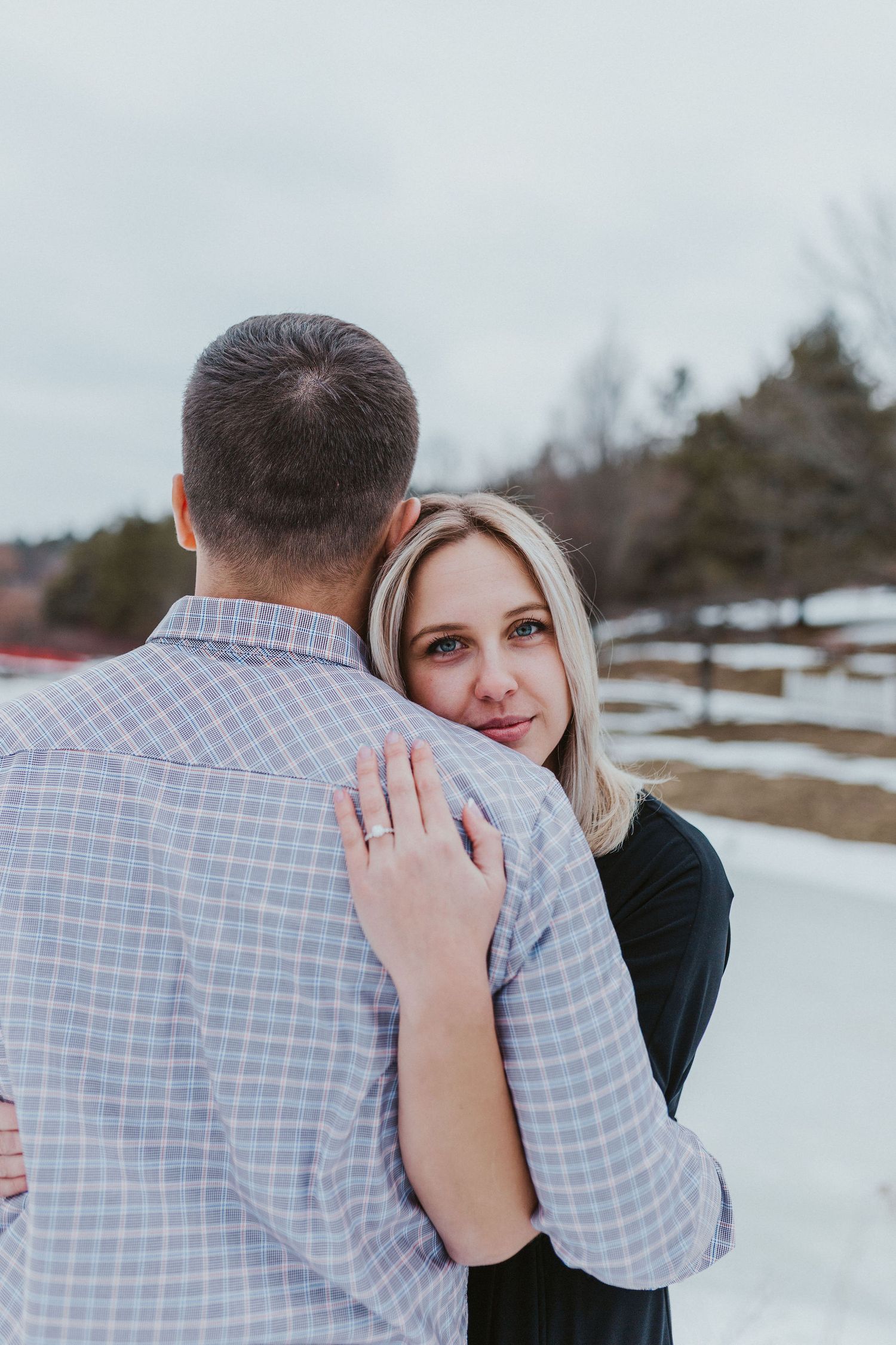 A blonde woman embraces a man in a plaid shirt outdoors in a snowy winter landscape.