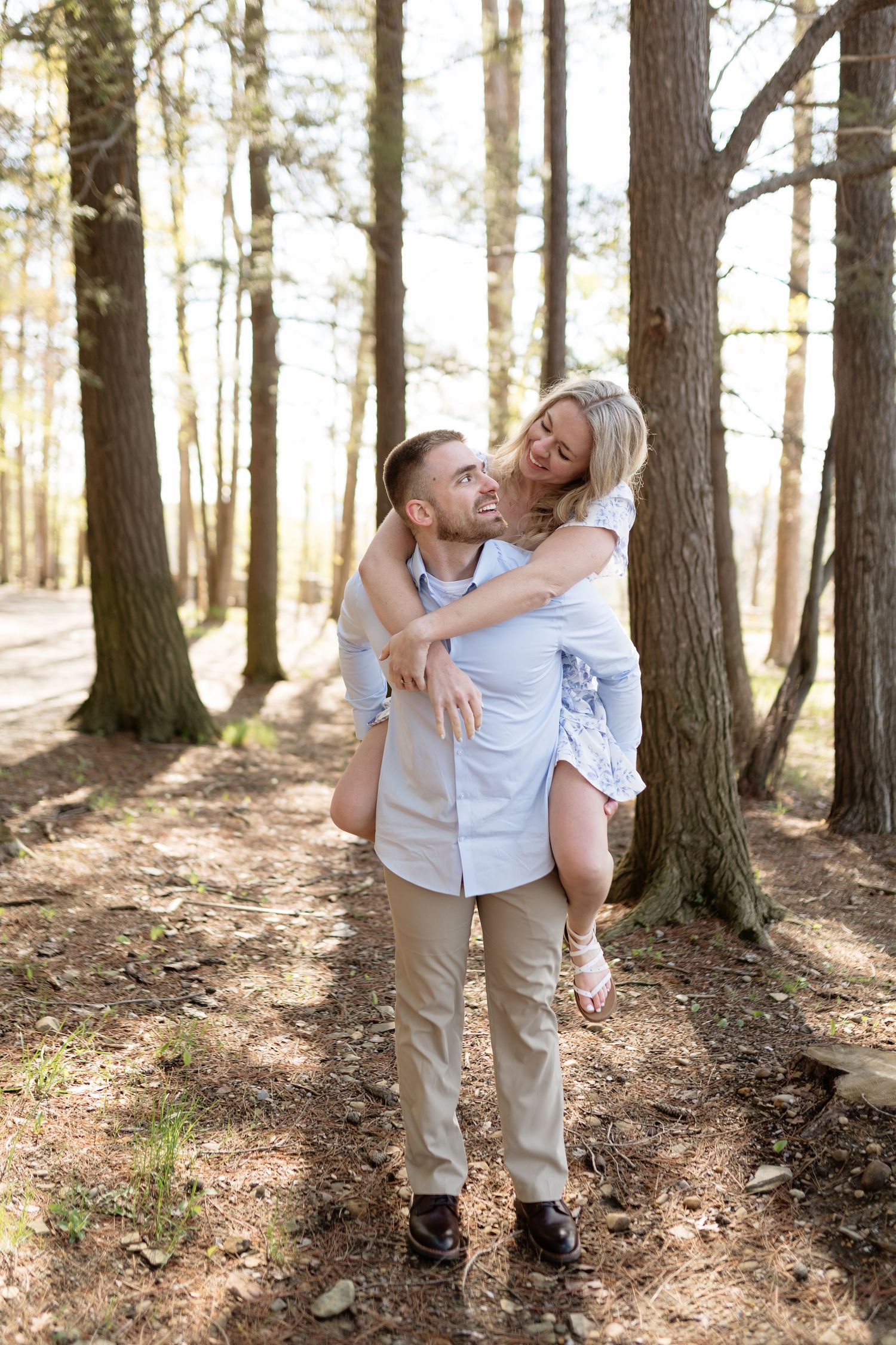 A happy couple enjoys a piggyback ride in a sunny pine forest, the woman laughing as the man carries her on his back.