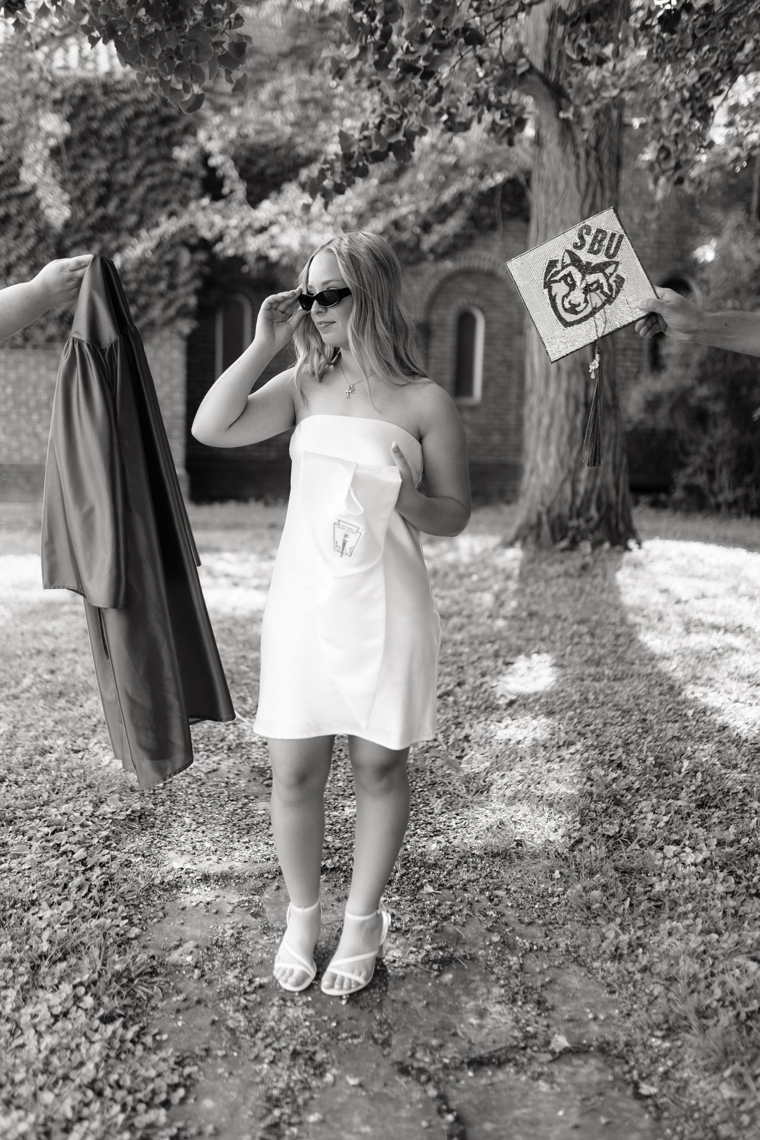 Young woman in white dress holding sunglasses with graduation gown and SBU sign in black and white outdoor photo.