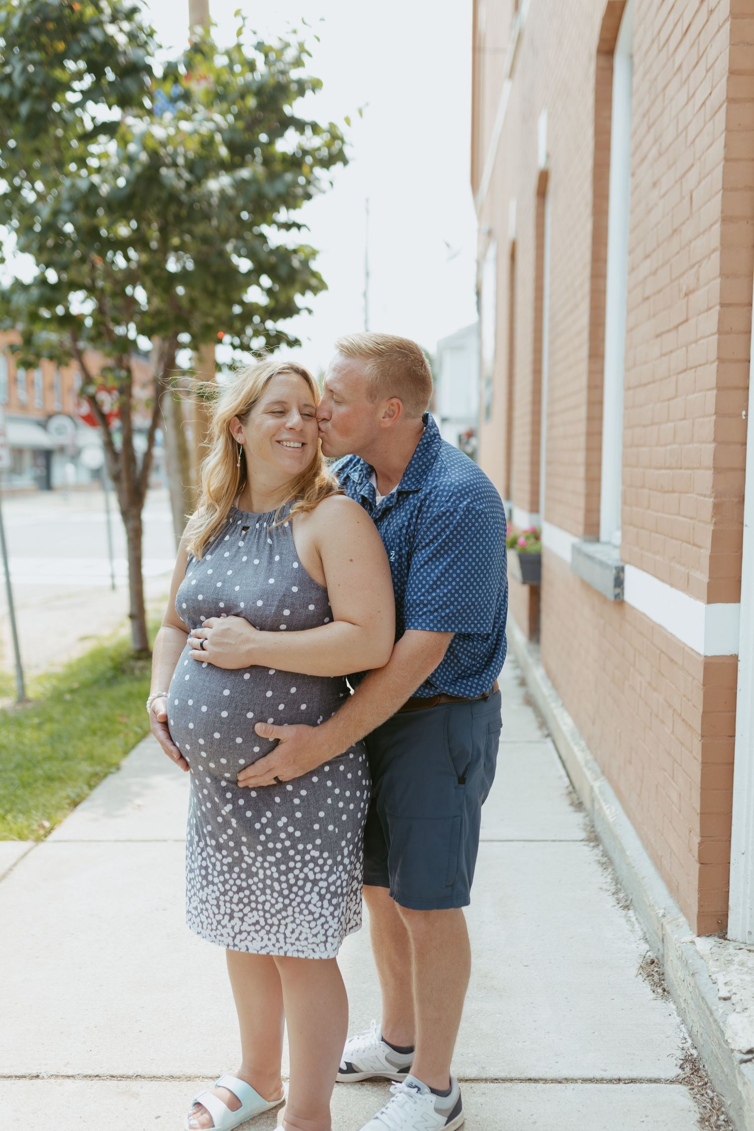 Pregnant woman in polka dot dress being kissed on cheek by man on sunny urban sidewalk beside brick building.