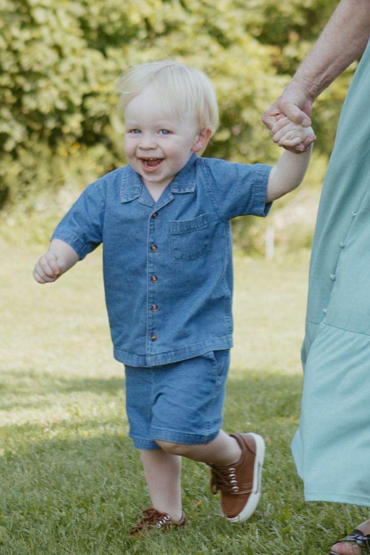 Smiling toddler boy in denim outfit holding adult hand while walking on green grass outdoors.