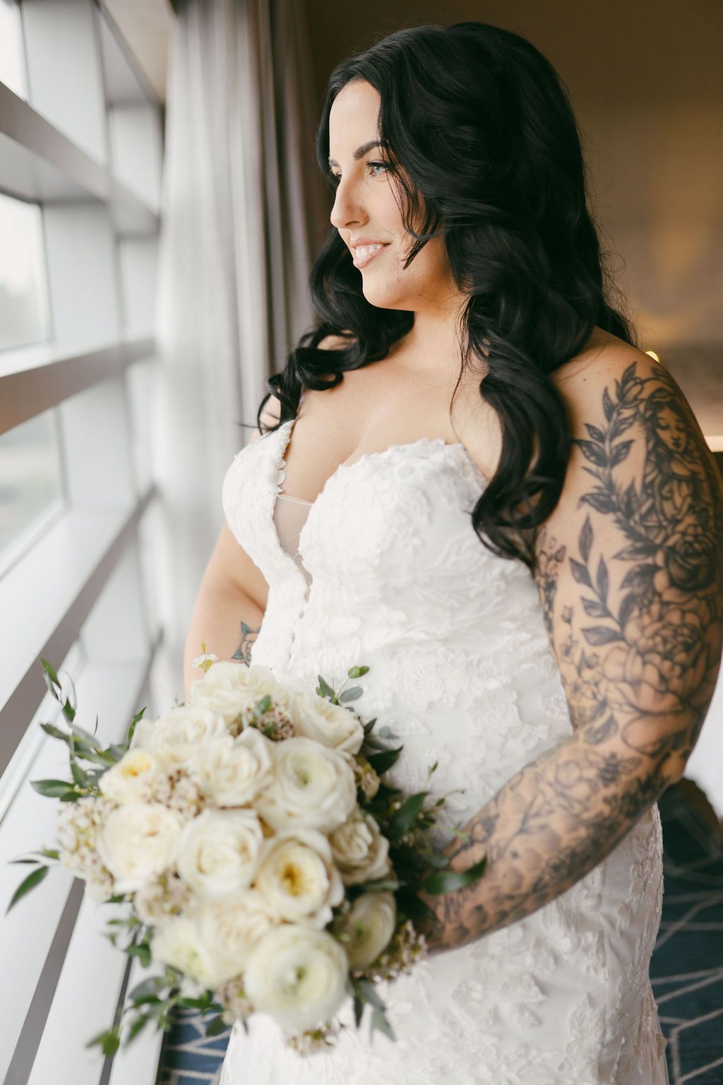 Tattooed bride in white lace gown holds white floral bouquet, smiling by window in soft natural light.