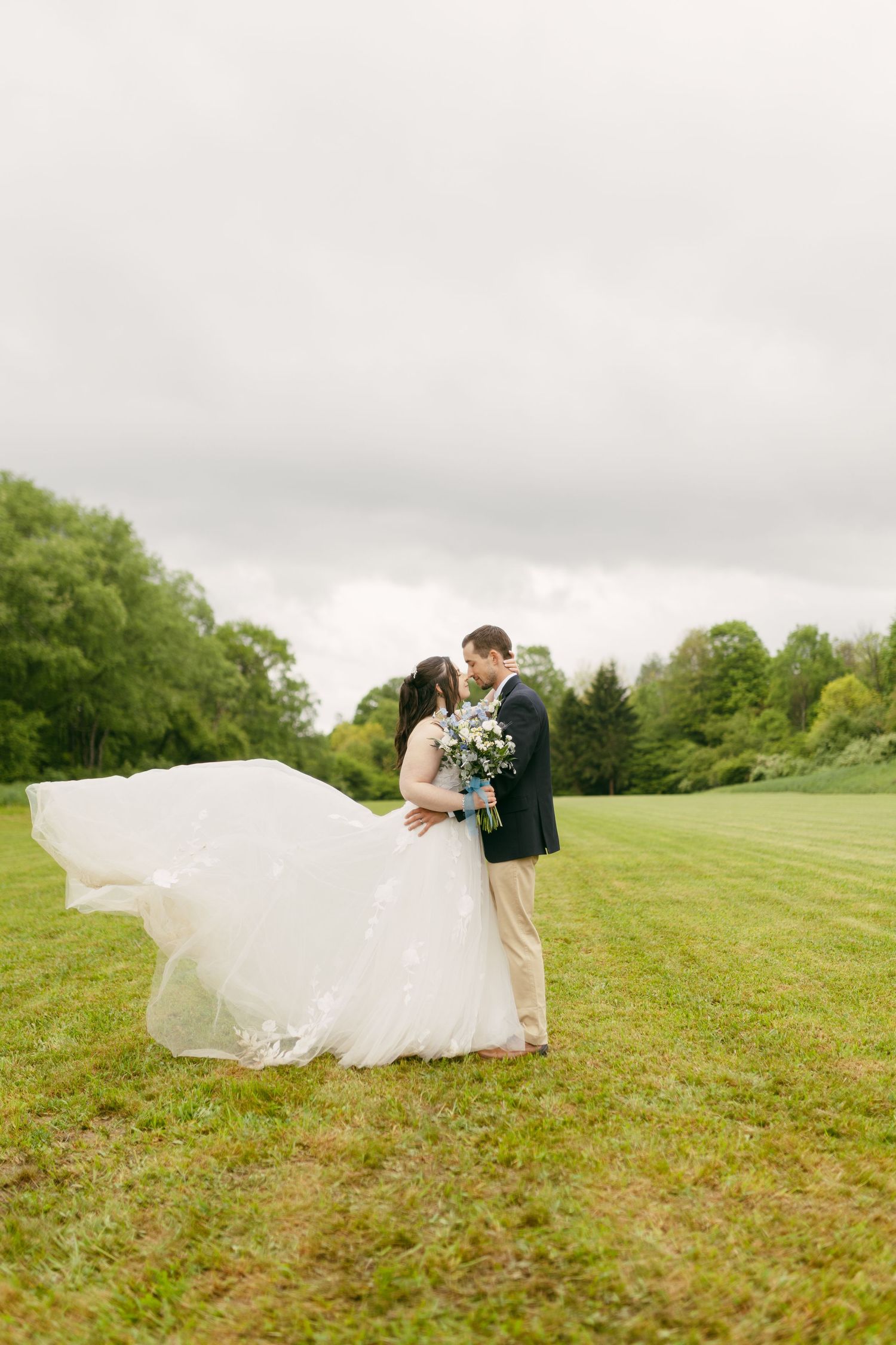 Bride and groom embracing on grassy hillside with flowing veil in the wind.