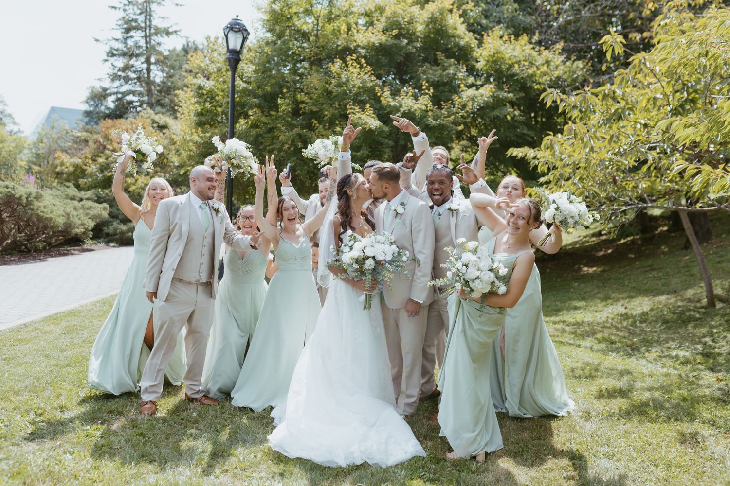 Wedding party celebrates outdoors, bride in white gown, bridesmaids in sage green dresses, groomsmen in tan suits, arms raised.