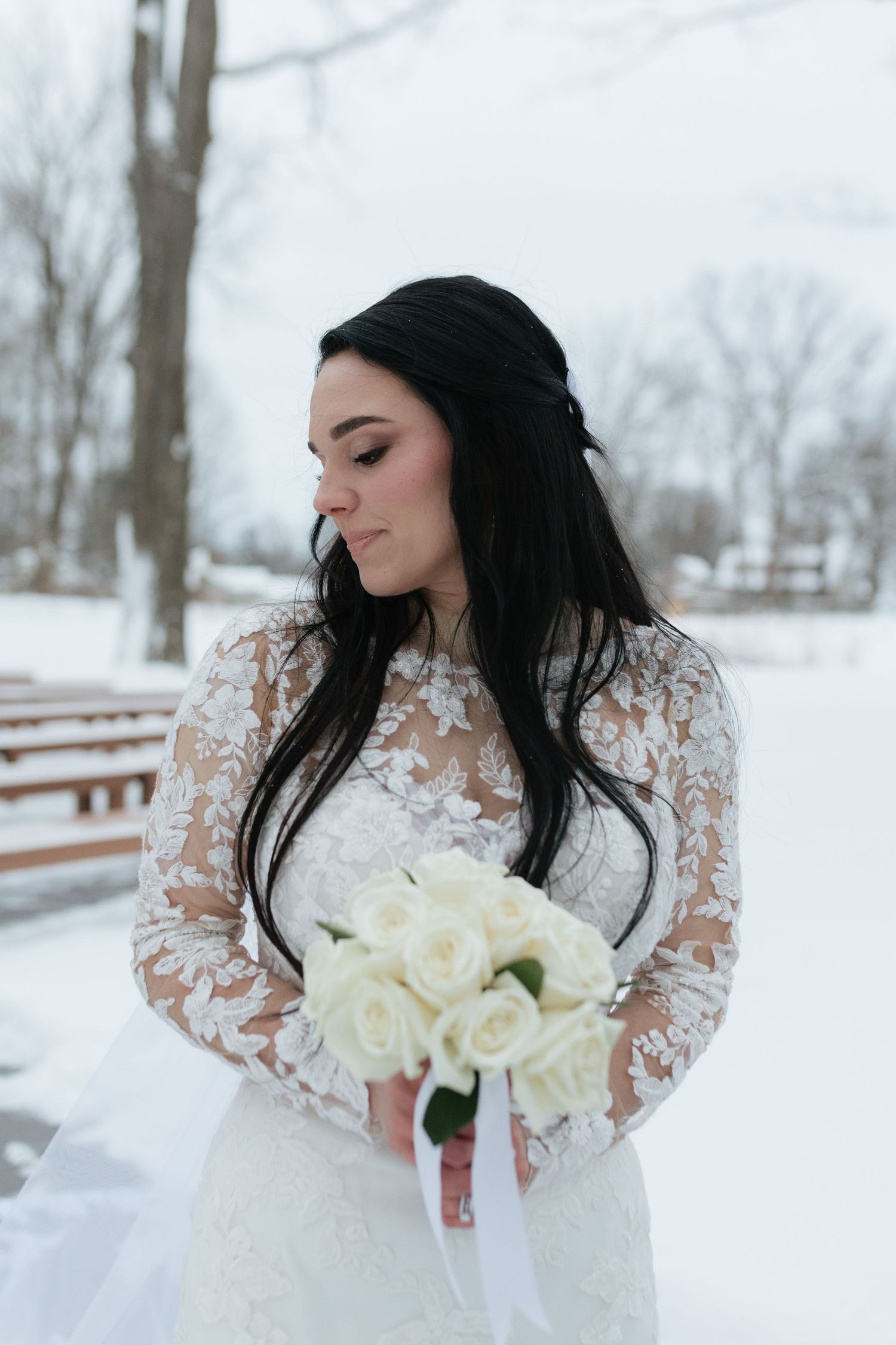 A bride in a lace wedding dress holds a bouquet of white roses against a snowy winter landscape.