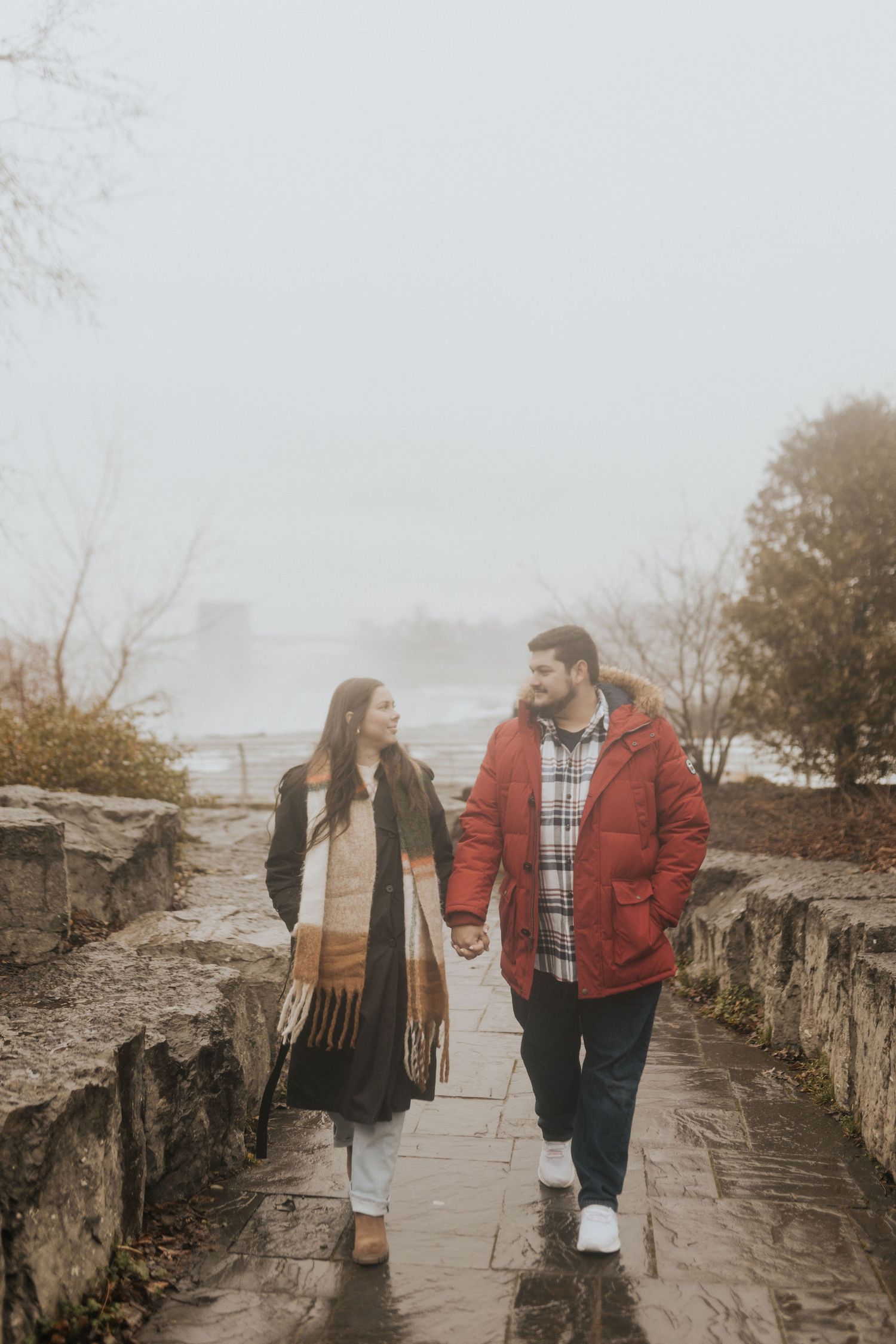 Couple holding hands walking on a foggy stone path near water, wearing winter coats and scarves.