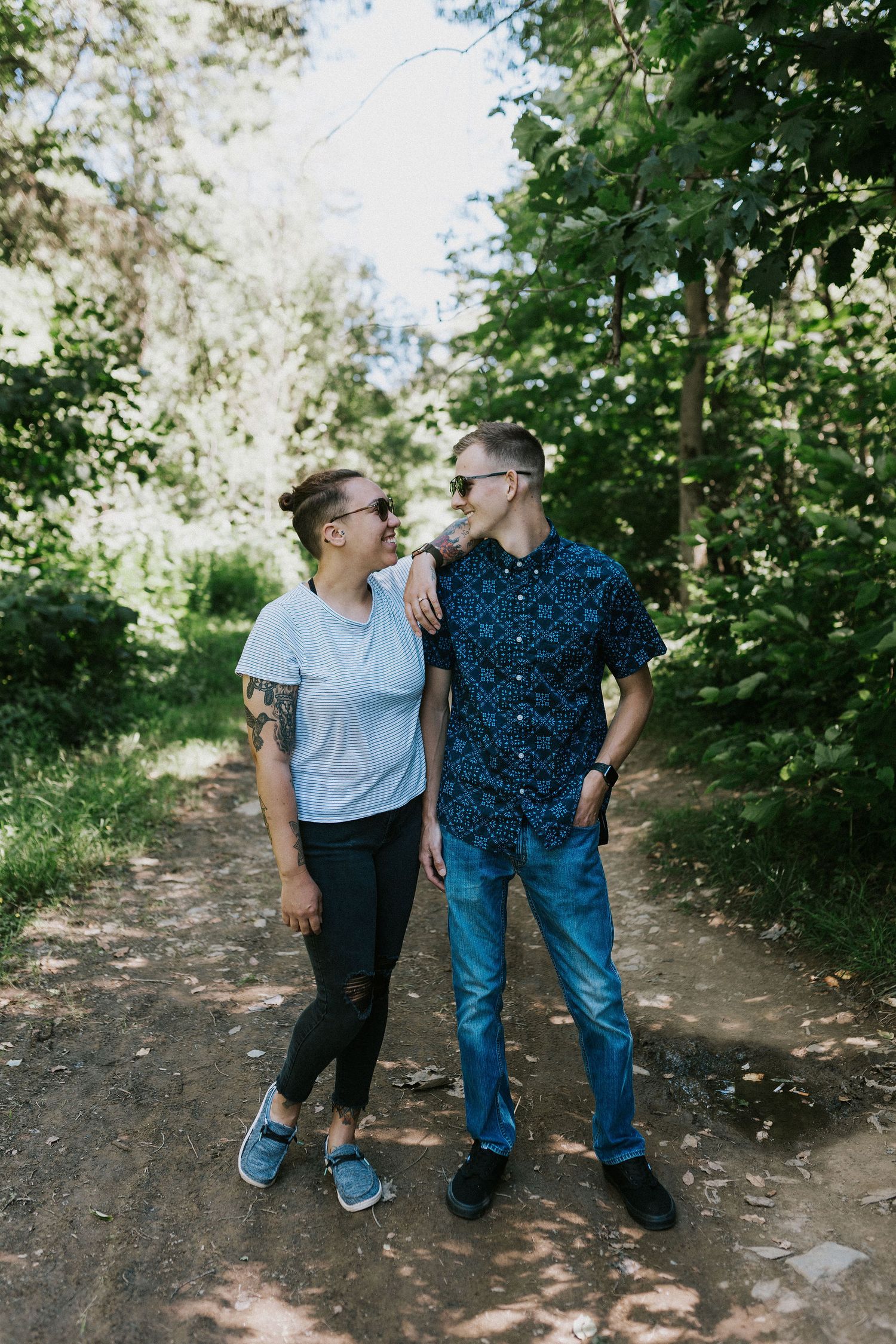 Couple walking on a forest trail, smiling at each other, wearing casual outfits and sunglasses on a sunny day.