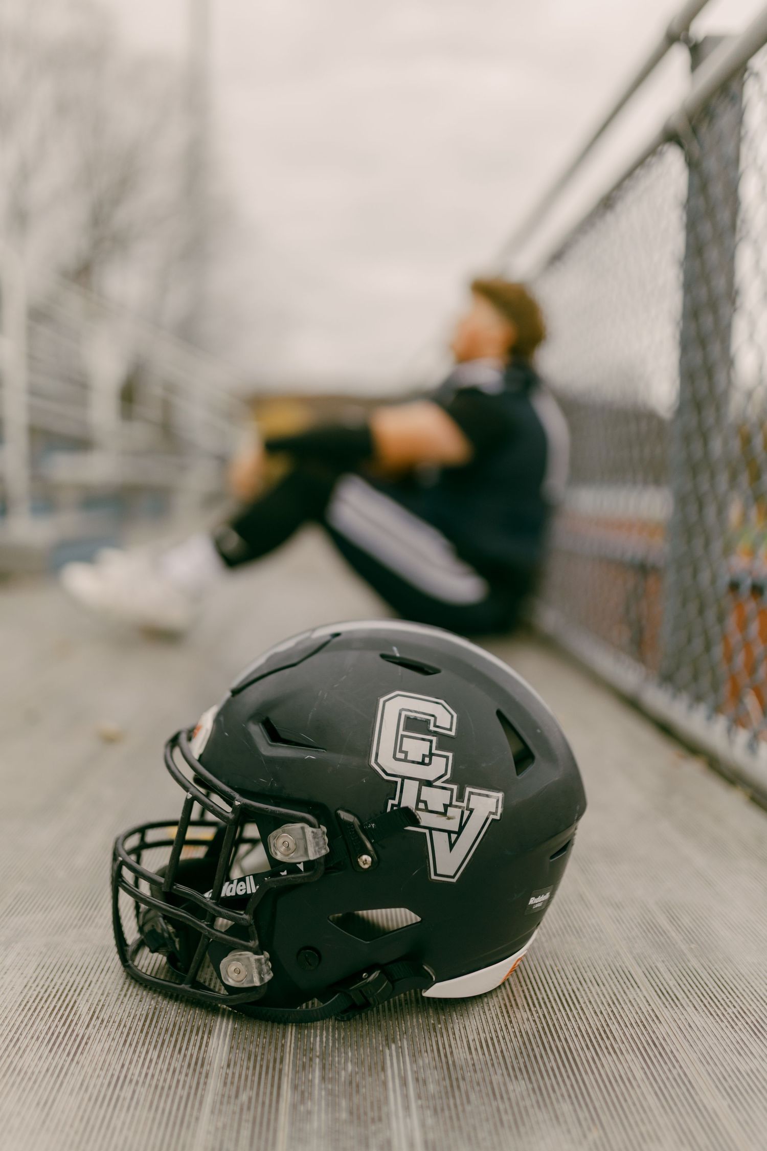 Black CV football helmet resting on bleachers with a player sitting against a chain-link fence in the blurred background.