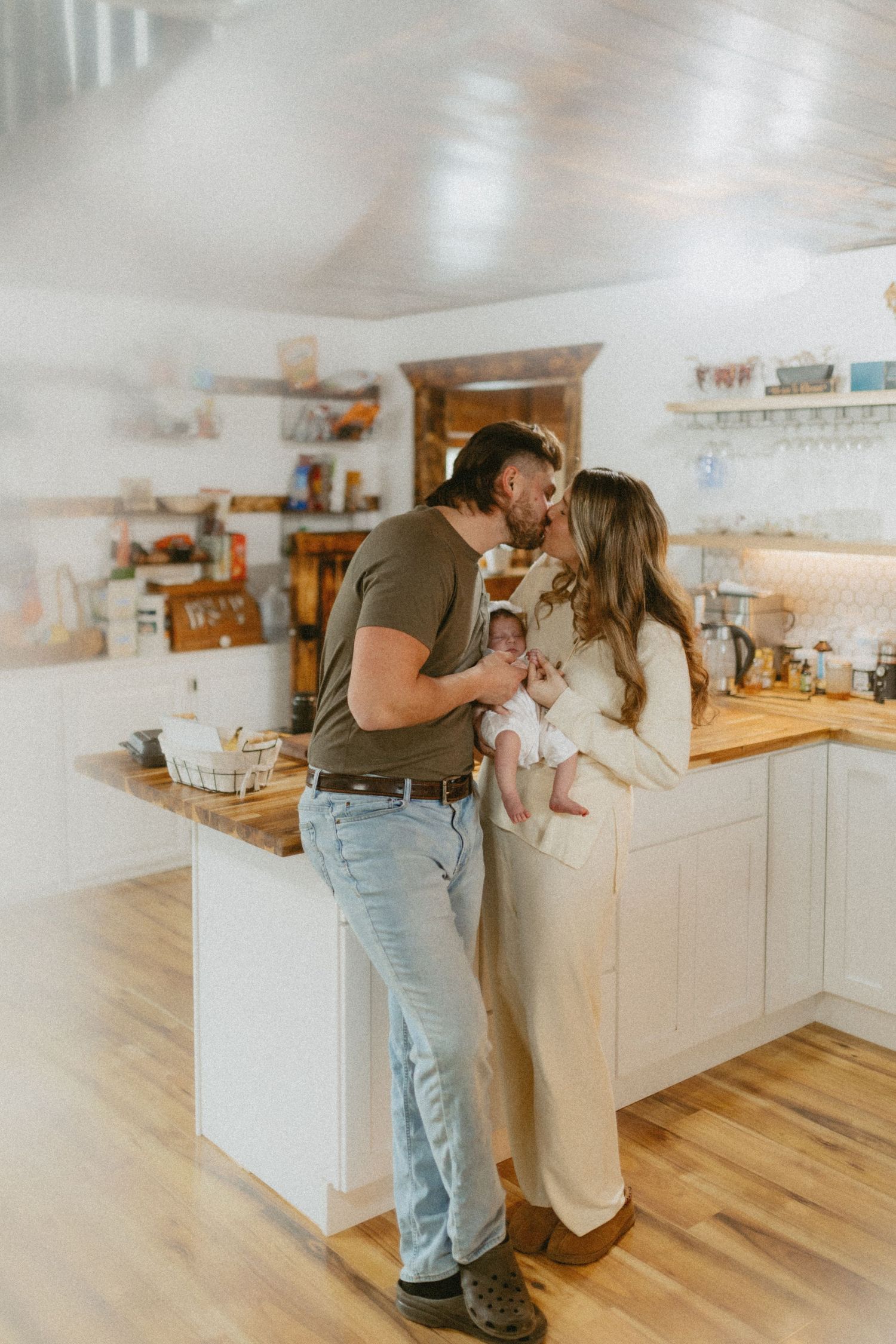 A couple shares a kiss while holding their newborn baby in a cozy white farmhouse kitchen.
