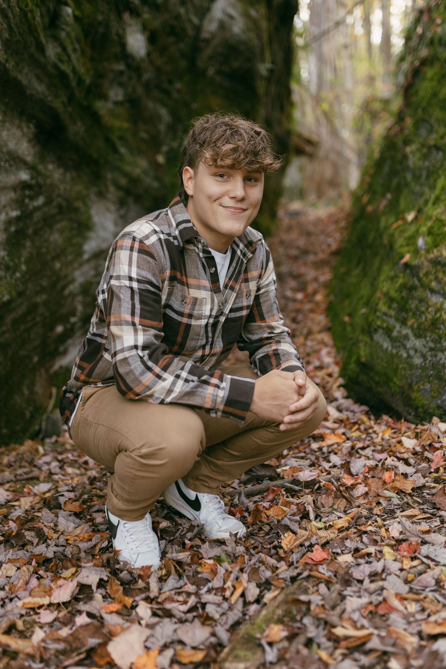Teen boy crouching among autumn leaves between mossy rocks, wearing plaid shirt and tan pants, smiling at camera.