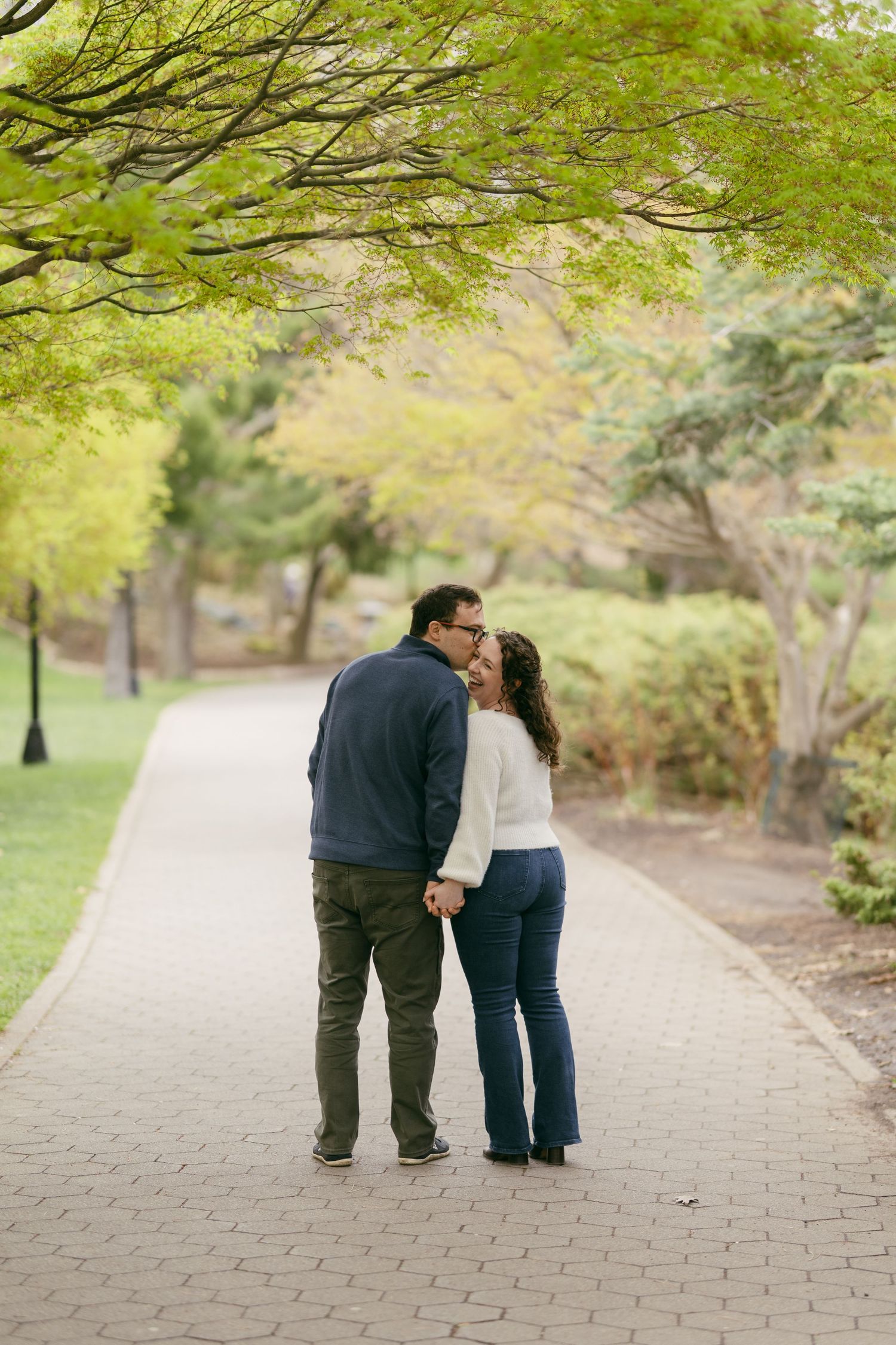 Couple sharing a kiss on a tree-lined brick pathway in a lush green park during spring.