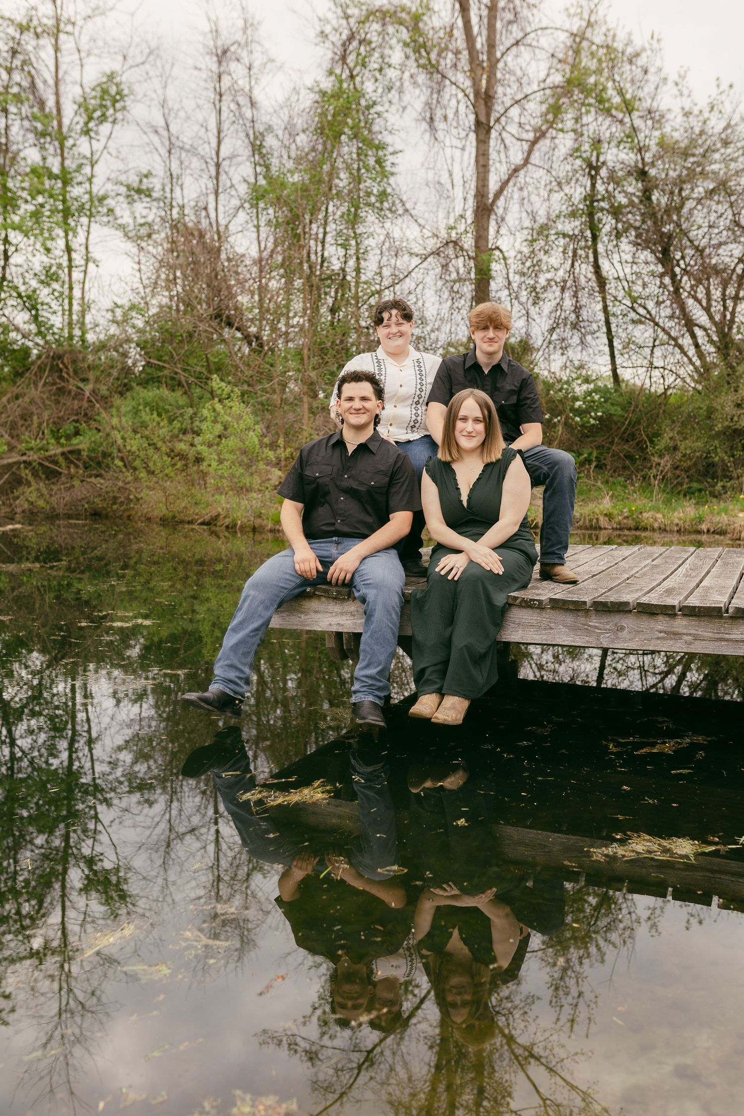 People sitting together on a wooden dock over a reflective pond surrounded by trees.