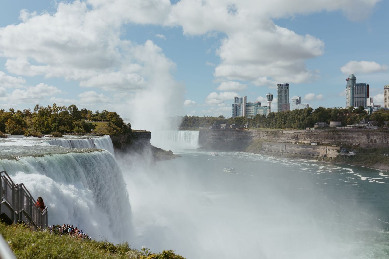 Aerial view of Niagara Falls with mist rising, lush greenery, and a city skyline visible in the background.