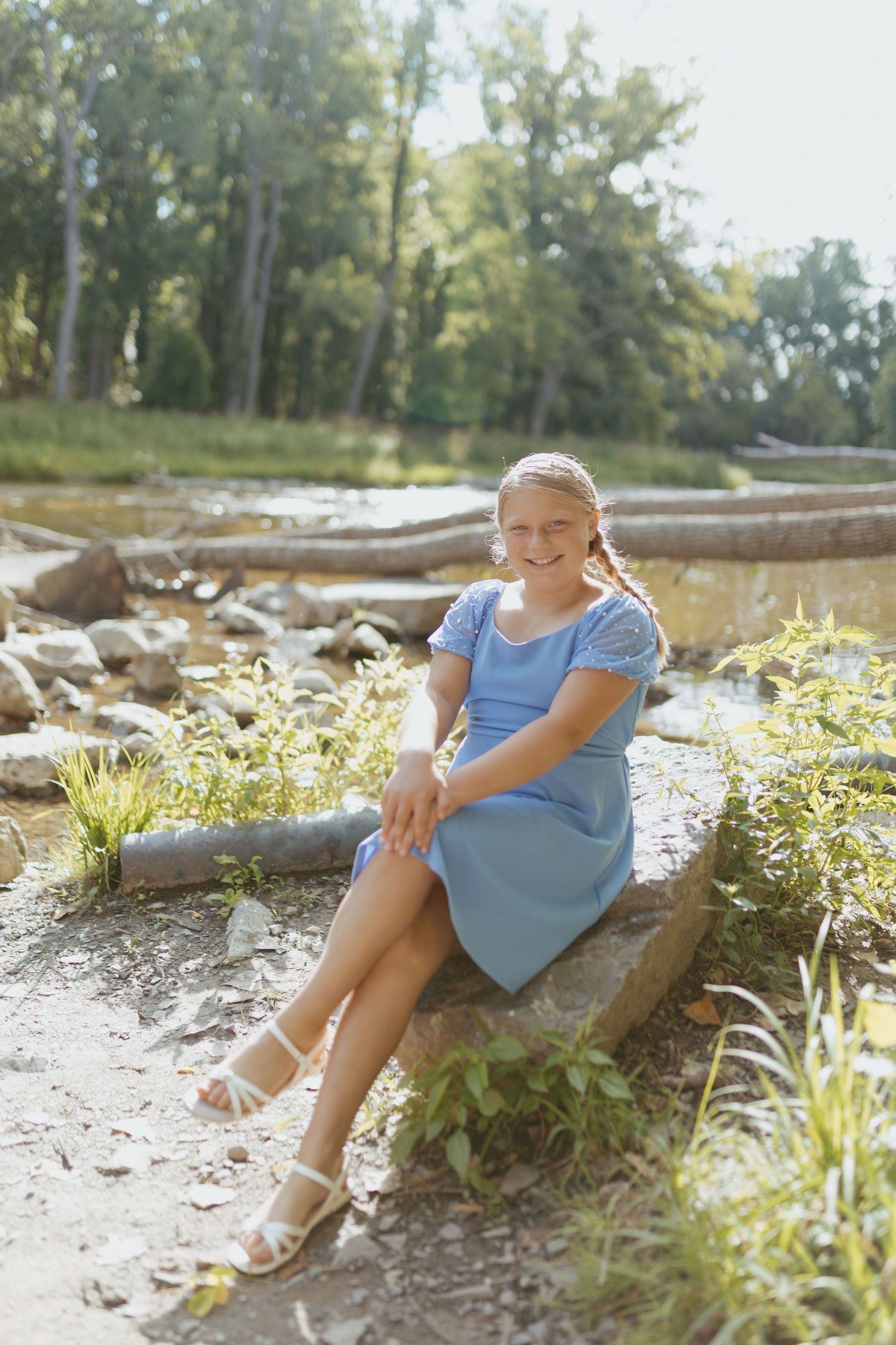 A smiling girl in a blue dress sits on a rock by a sunny riverside surrounded by lush green trees.