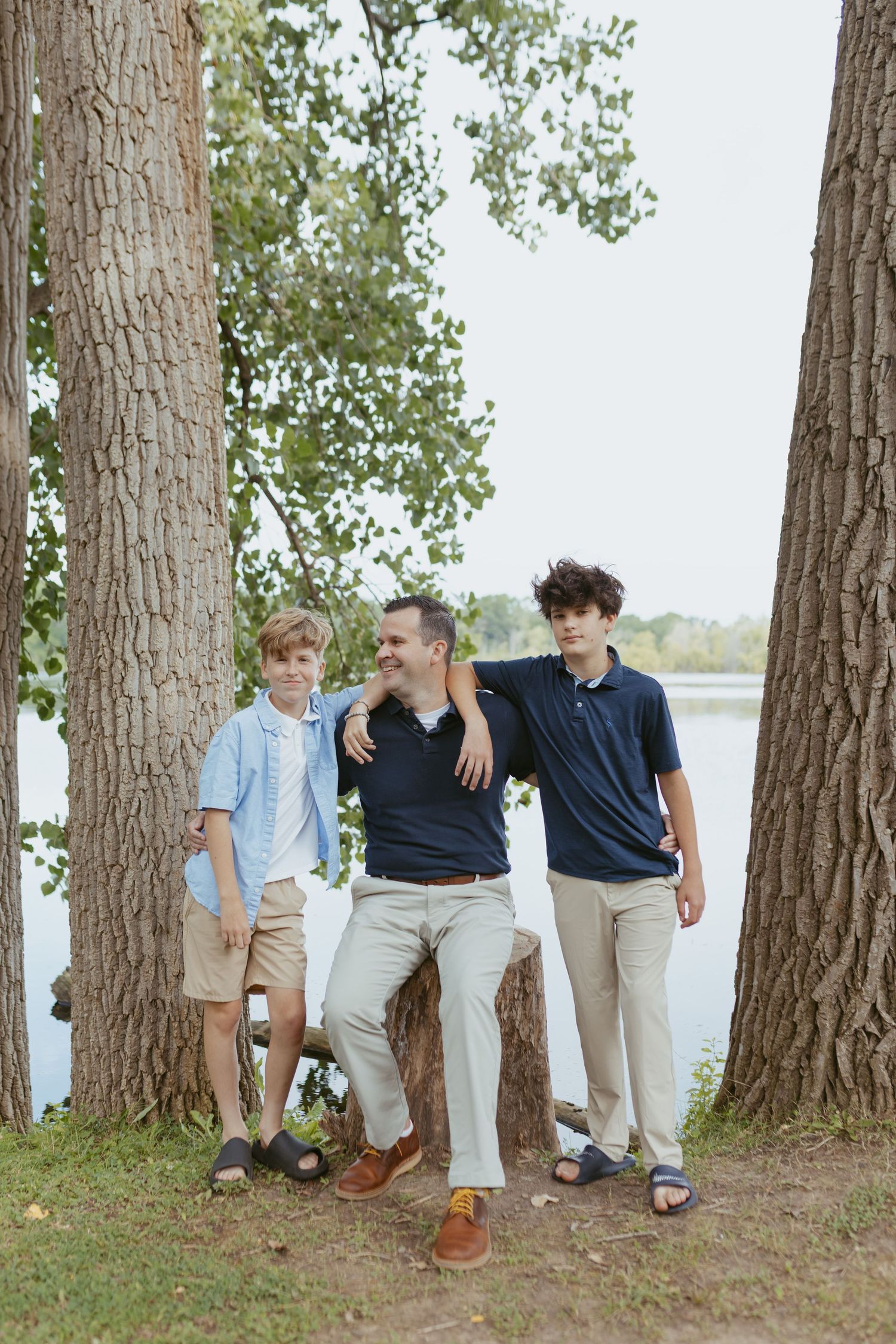 A father and two sons pose together near trees by a lake in a warm outdoor family portrait.