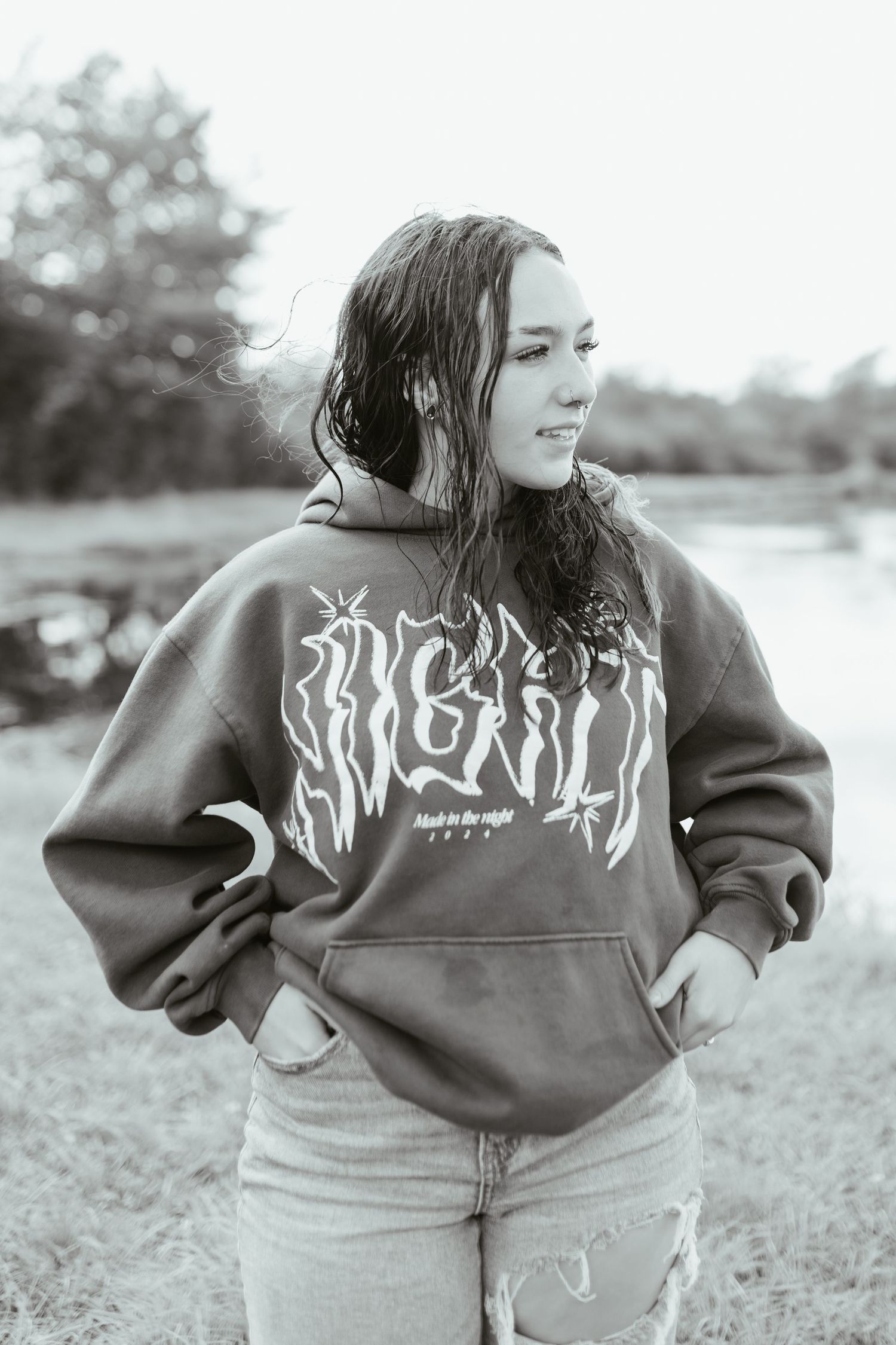 Black and white photo of a young woman in an oversized graphic hoodie and jeans, posing outdoors near water.